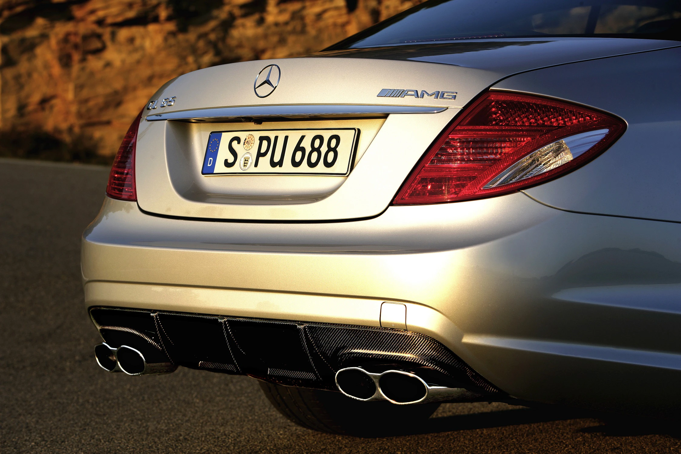 Rückansicht eines silbernen Mercedes AMG mit vier Auspuffrohren, geparkt auf einer Straße vor einer felsigen Kulisse bei Sonnenuntergang.