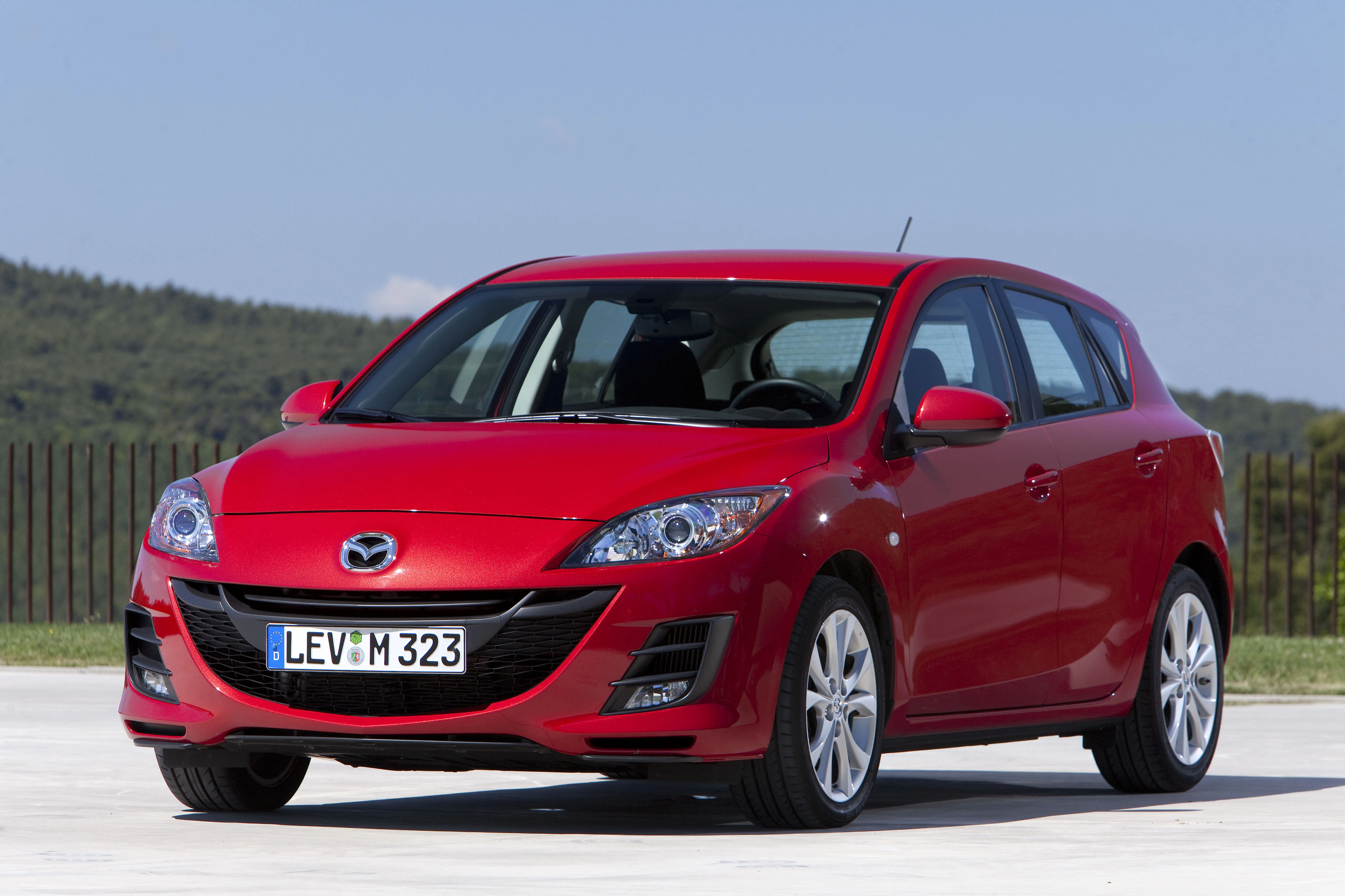 Ein roter Mazda 3 Kompaktwagen steht auf einem Parkplatz vor einer grünen Landschaft bei klarem Himmel.