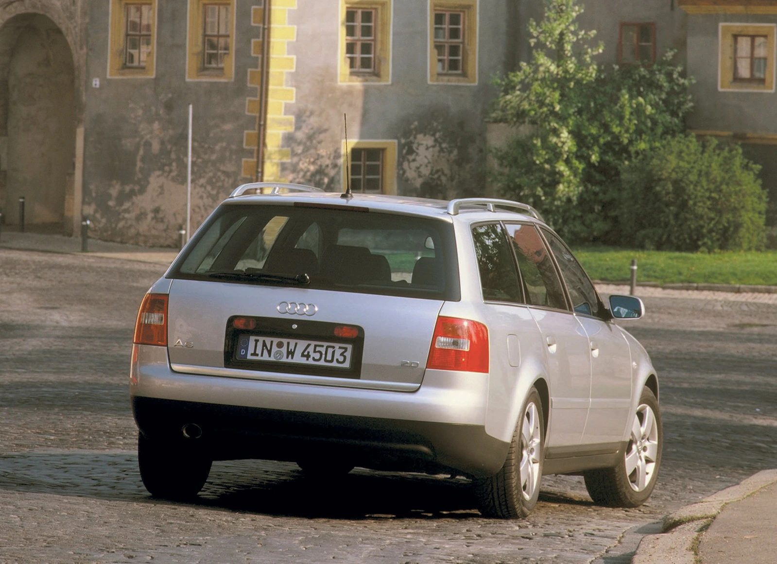 Silberner Audi Kombi auf Kopfsteinpflaster vor historischem Gebäude mit gelber Umrandung der Fenster und grünen Pflanzen im Hintergrund.