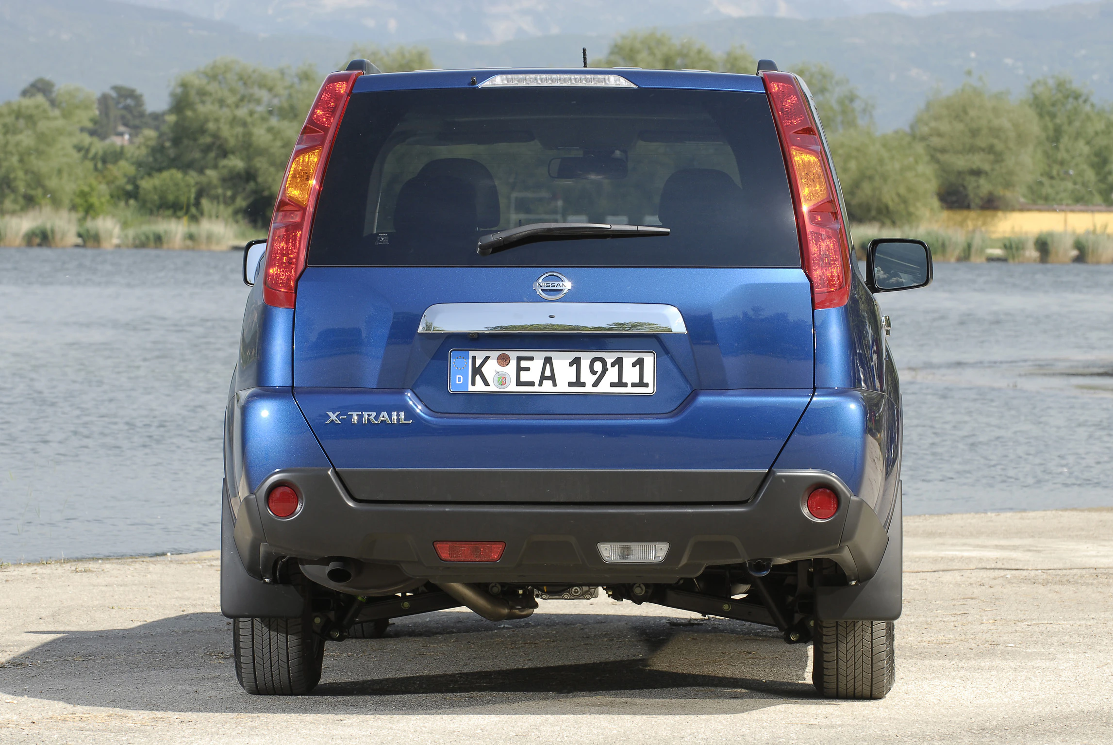 Blauer Nissan X-Trail parkt rückwärts vor einem See mit Bäumen und Bergen im Hintergrund. Autokennzeichen: K EA 1911.