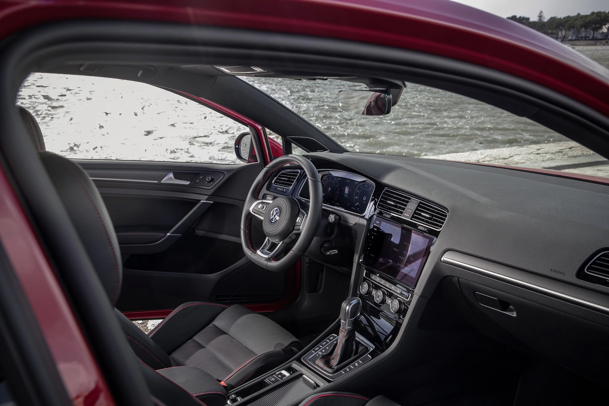 Innenraum eines roten Autos mit modernen Armaturenbrett, Lenkrad, Schalthebel und Touchscreen-Display. Blick auf das Wasser draußen.