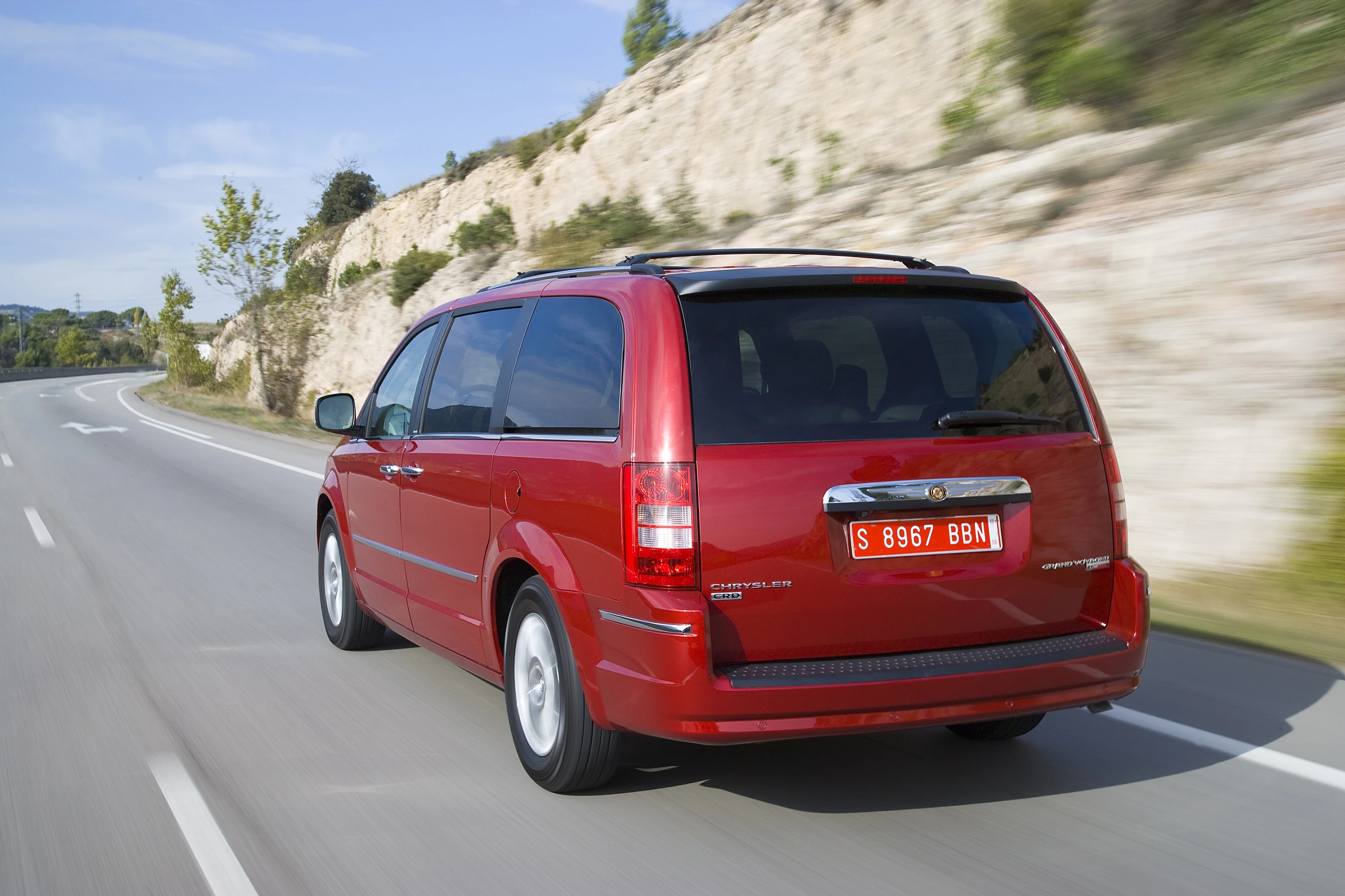 Roter Minivan fährt auf einer Landstraße entlang einer steinigen Landschaft. Lizenzschild mit Nummer „S 8967 BBN“.