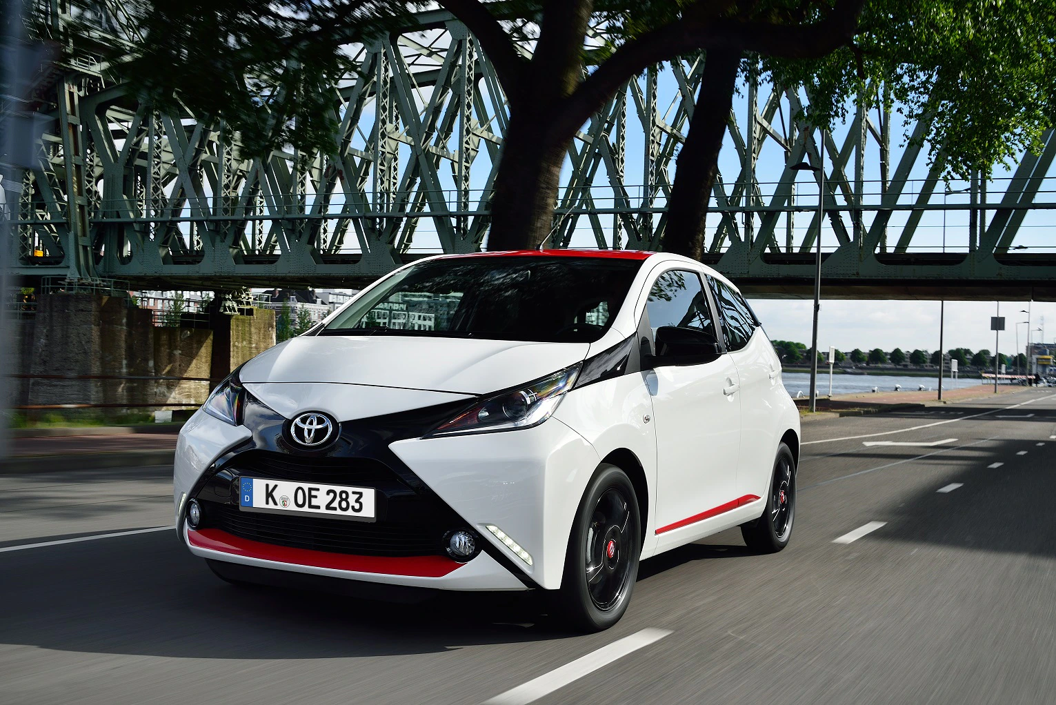 Ein weißer Toyota Aygo fährt auf einer Straße neben einem Fluss. Im Hintergrund ist eine Brücke und Bäume zu sehen.