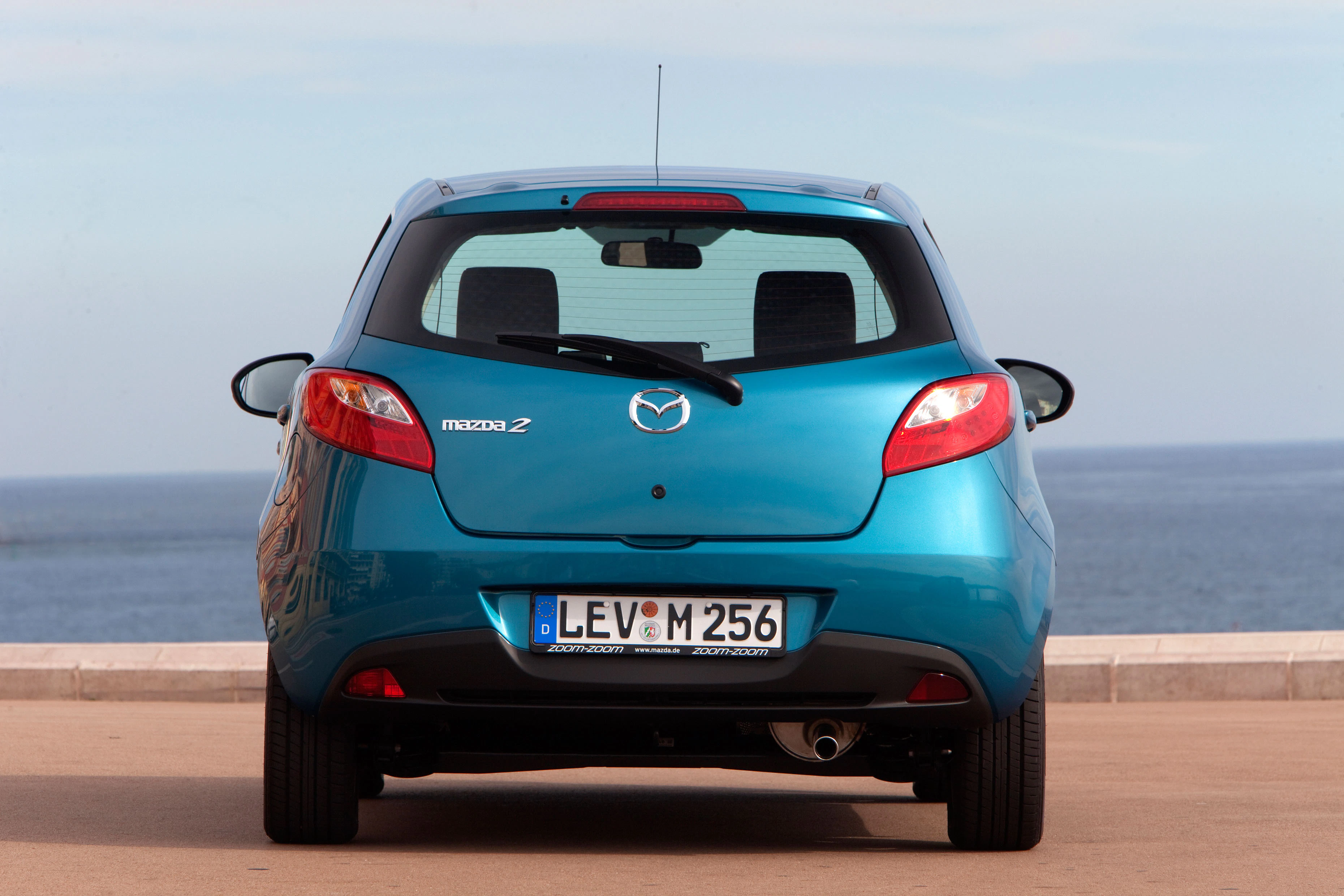 Blauer Mazda 2 von hinten, geparkt vor einem Meereshintergrund. Europäisches Nummernschild sichtbar.
