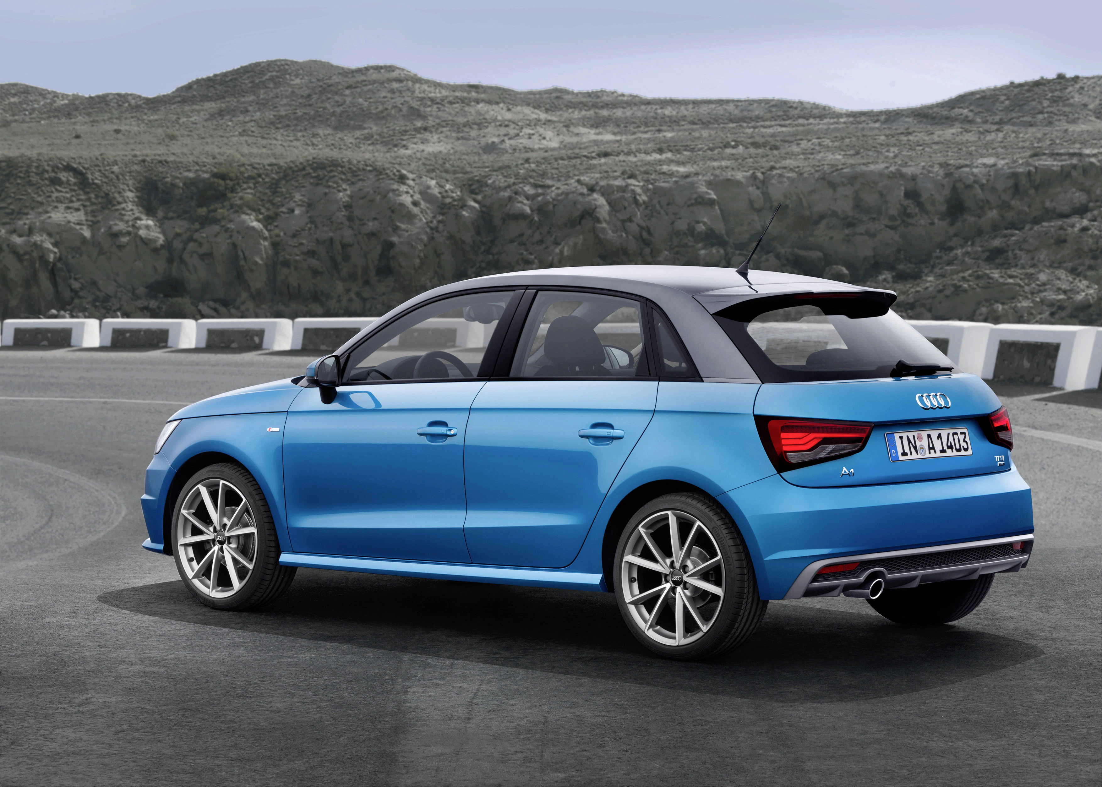 Ein blauer Audi A1 Sportback parkt auf einer asphaltierten Fläche vor einer grünen Felslandschaft. Die Rückseite des Fahrzeugs ist sichtbar.