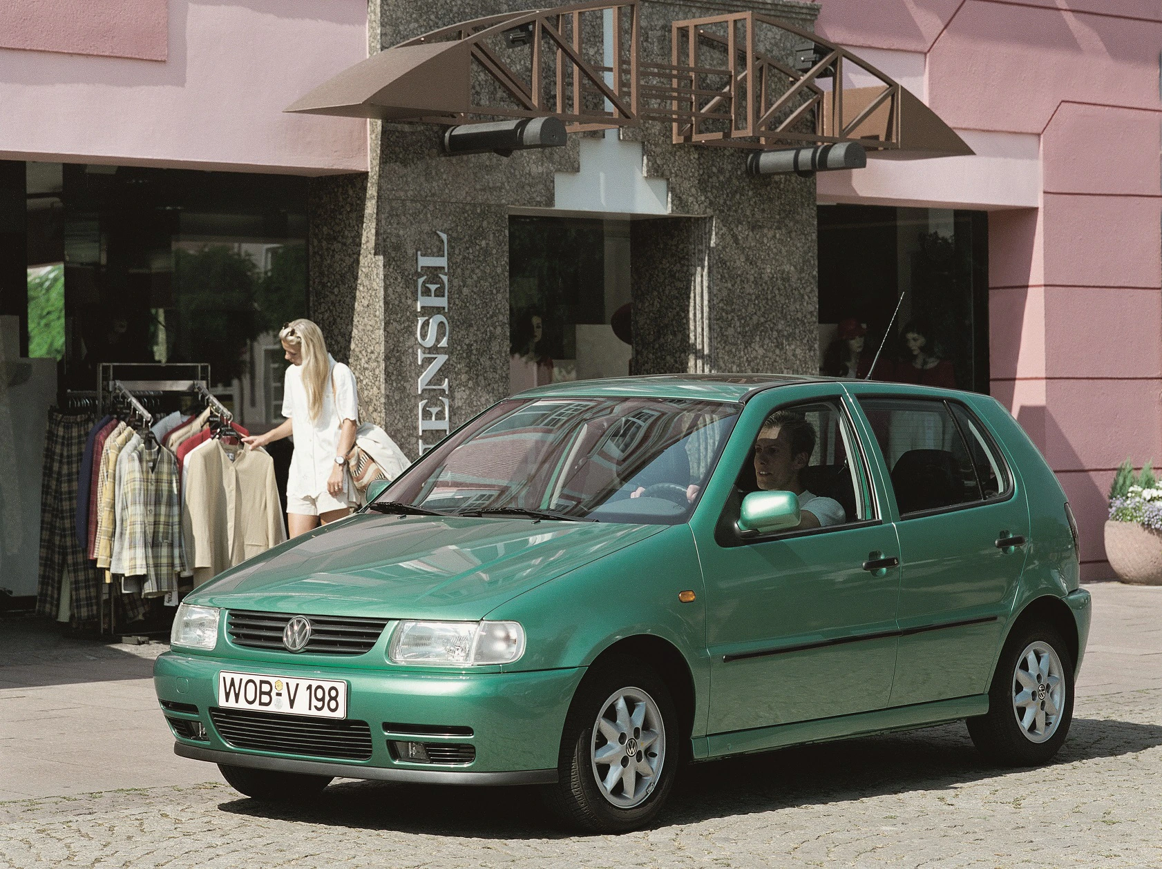 Grüner VW Polo parkt vor einem Bekleidungsgeschäft, Fußgänger im Hintergrund, sonniger Tag.