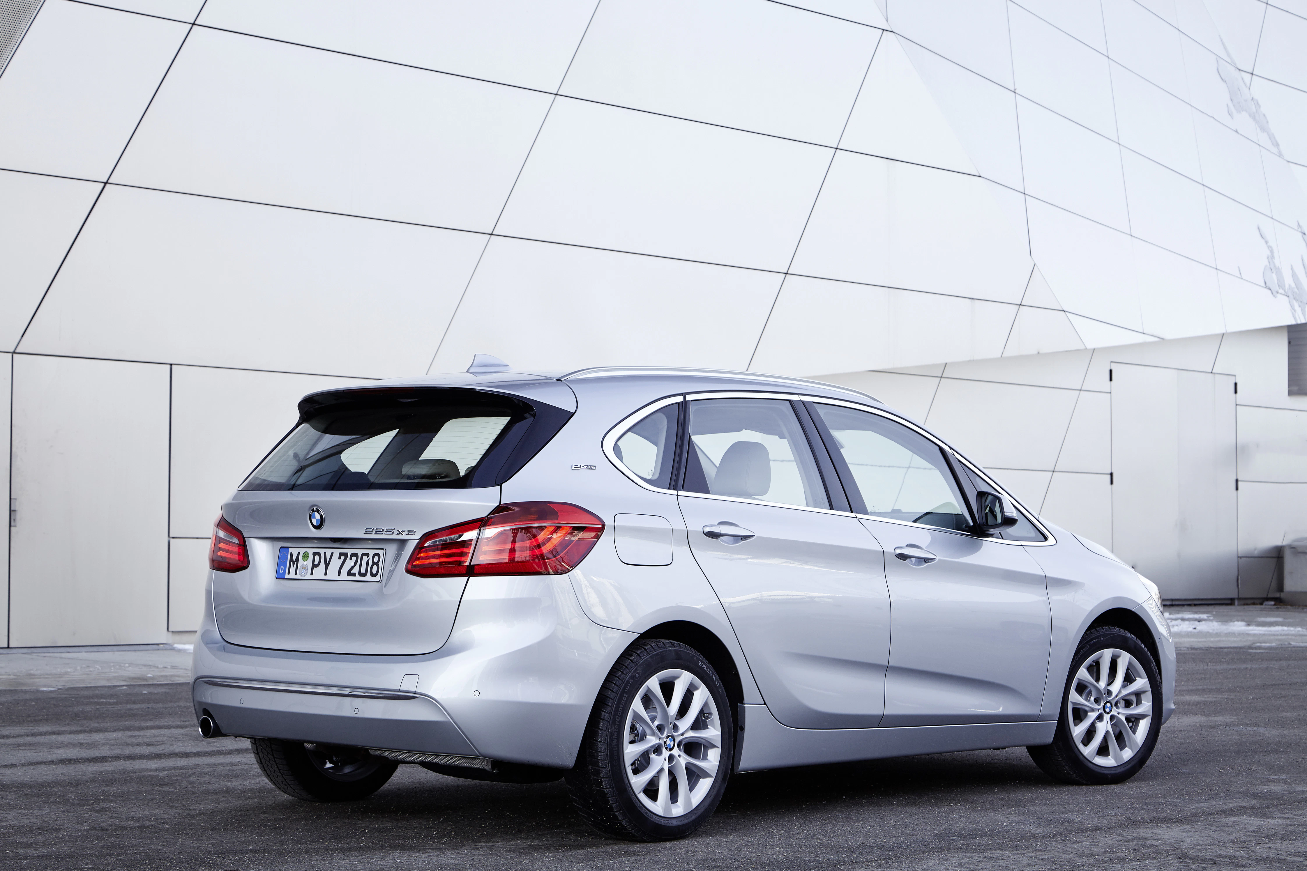 Silberner BMW 2er Active Tourer geparkt vor moderner, weißer Gebäudewand. Rechte Seitenansicht des Autos, geländetypische Umgebung.