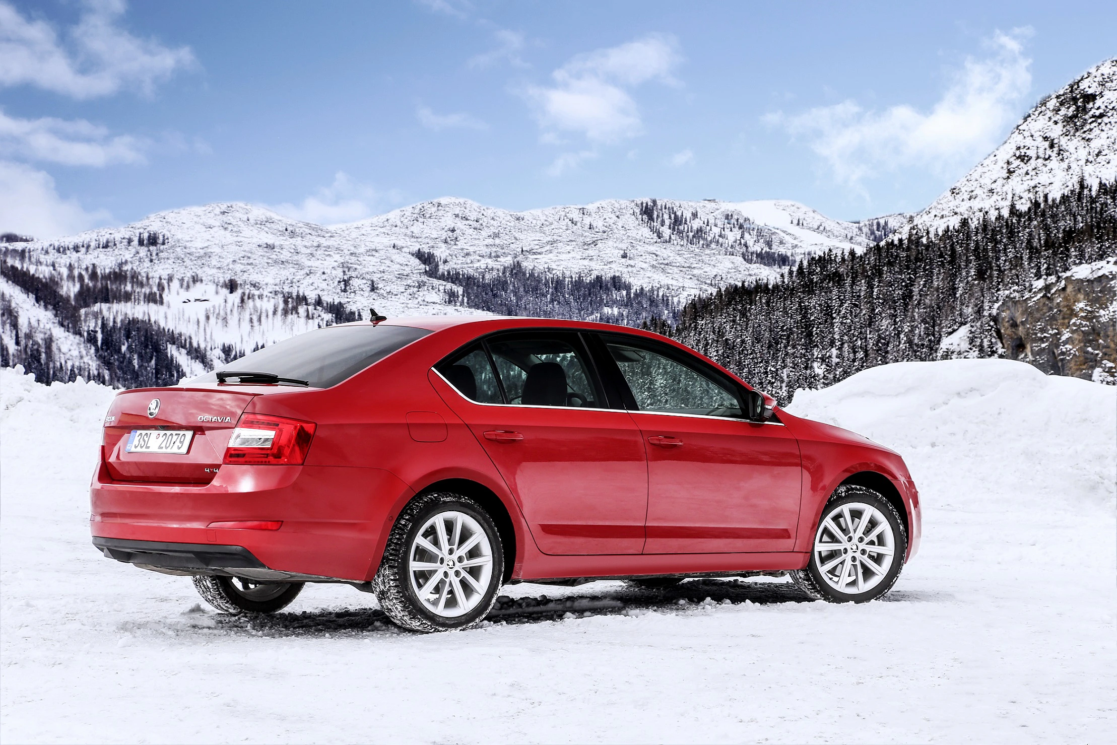 Rotes Auto parkt auf schneebedecktem Gelände, umgeben von schneebedeckten Bergen und Bäumen unter blauem Himmel.