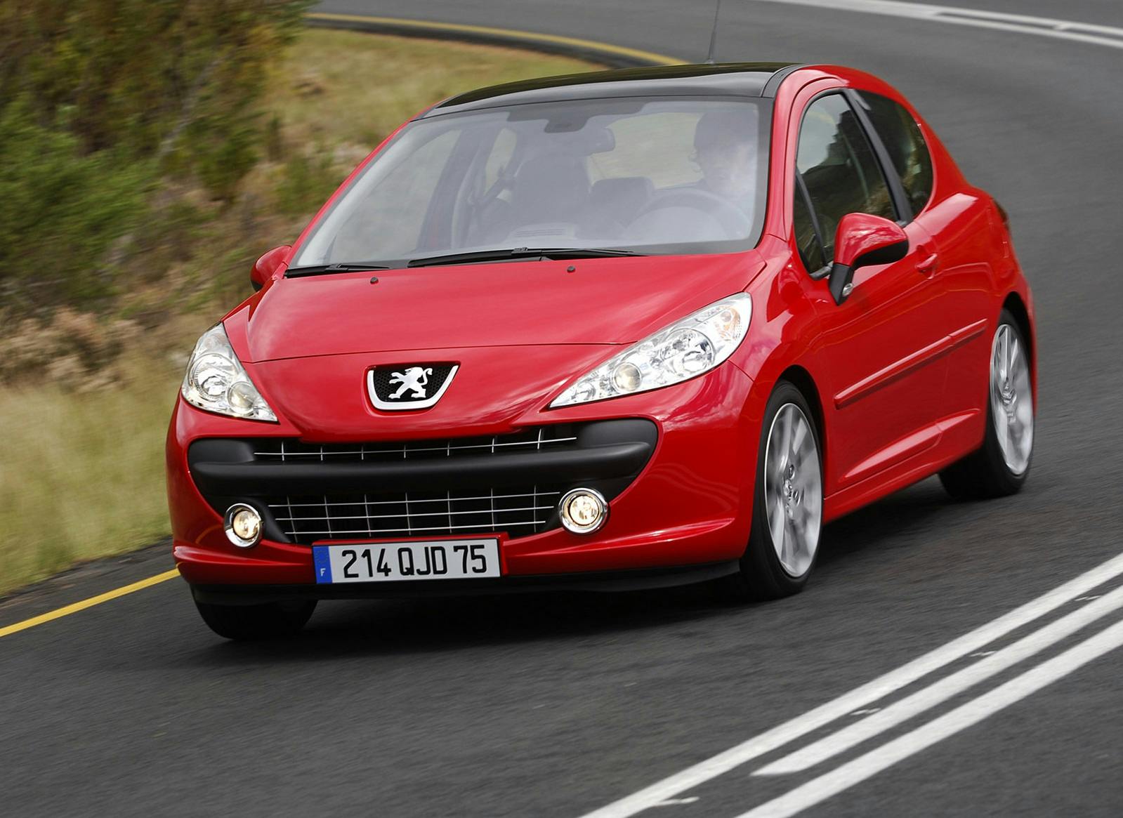 Roter Peugeot 207 fährt auf kurviger Landstraße. Auto mit runden Scheinwerfern und französischem Kennzeichen in grüner Landschaft.