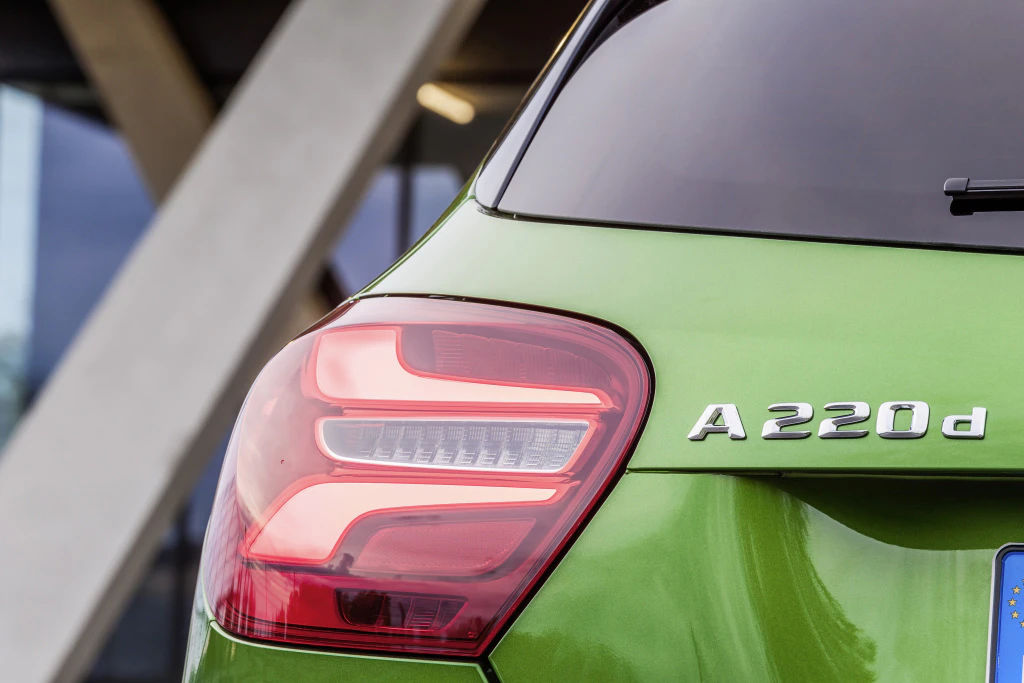 Grüner Mercedes A220d von hinten, zeigt Rücklicht und Emblem im Fokus. Modernes Design mit architektonischem Hintergrund.