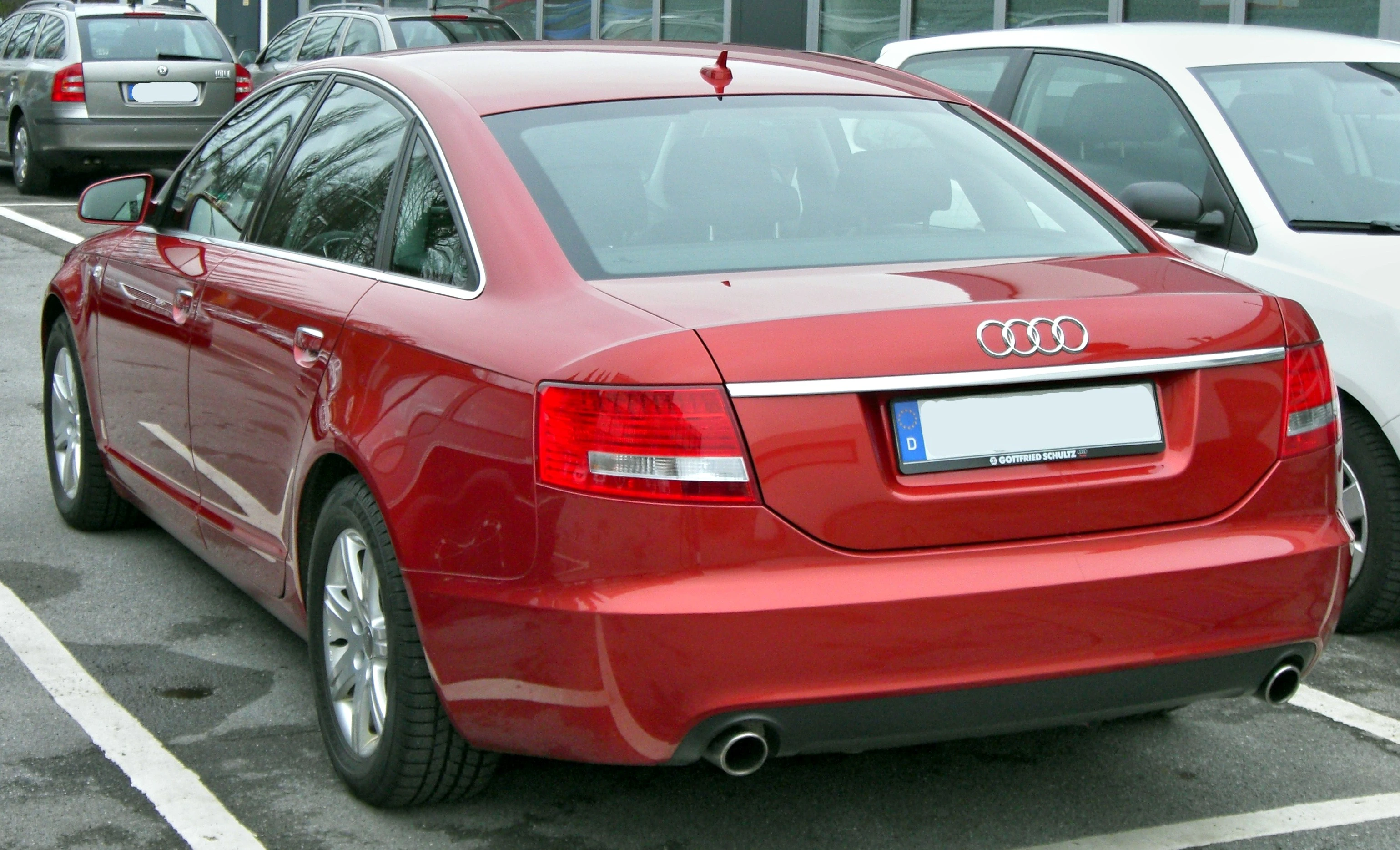 Roter Audi A6, geparkt auf einem Parkplatz, Heckansicht, im Hintergrund weitere geparkte Autos und ein Gebäudeteil.
