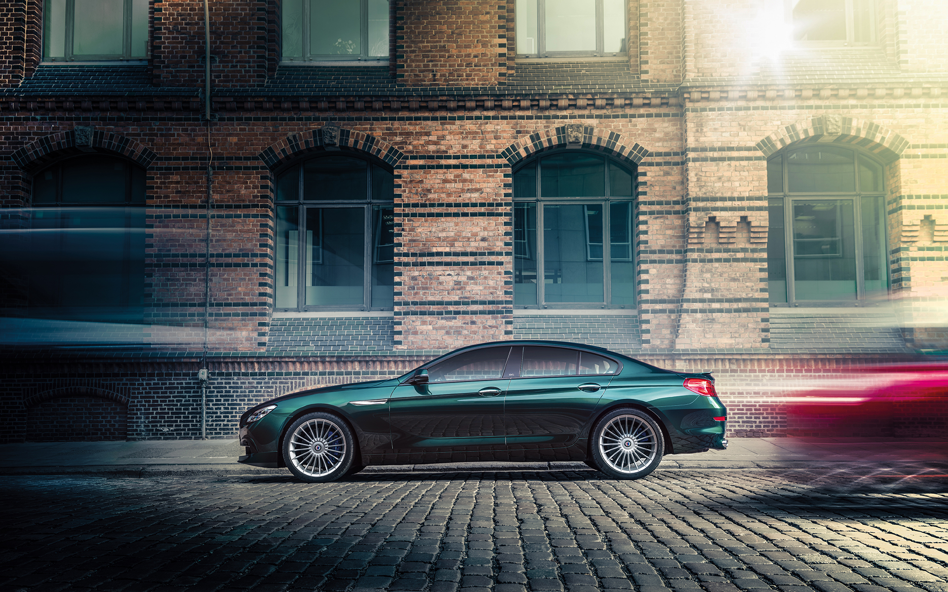Grünes Auto parkt auf einer Kopfsteinpflasterstraße vor einer roten Backsteinfassade, während Lichter von vorbeifahrenden Fahrzeugen erscheinen.