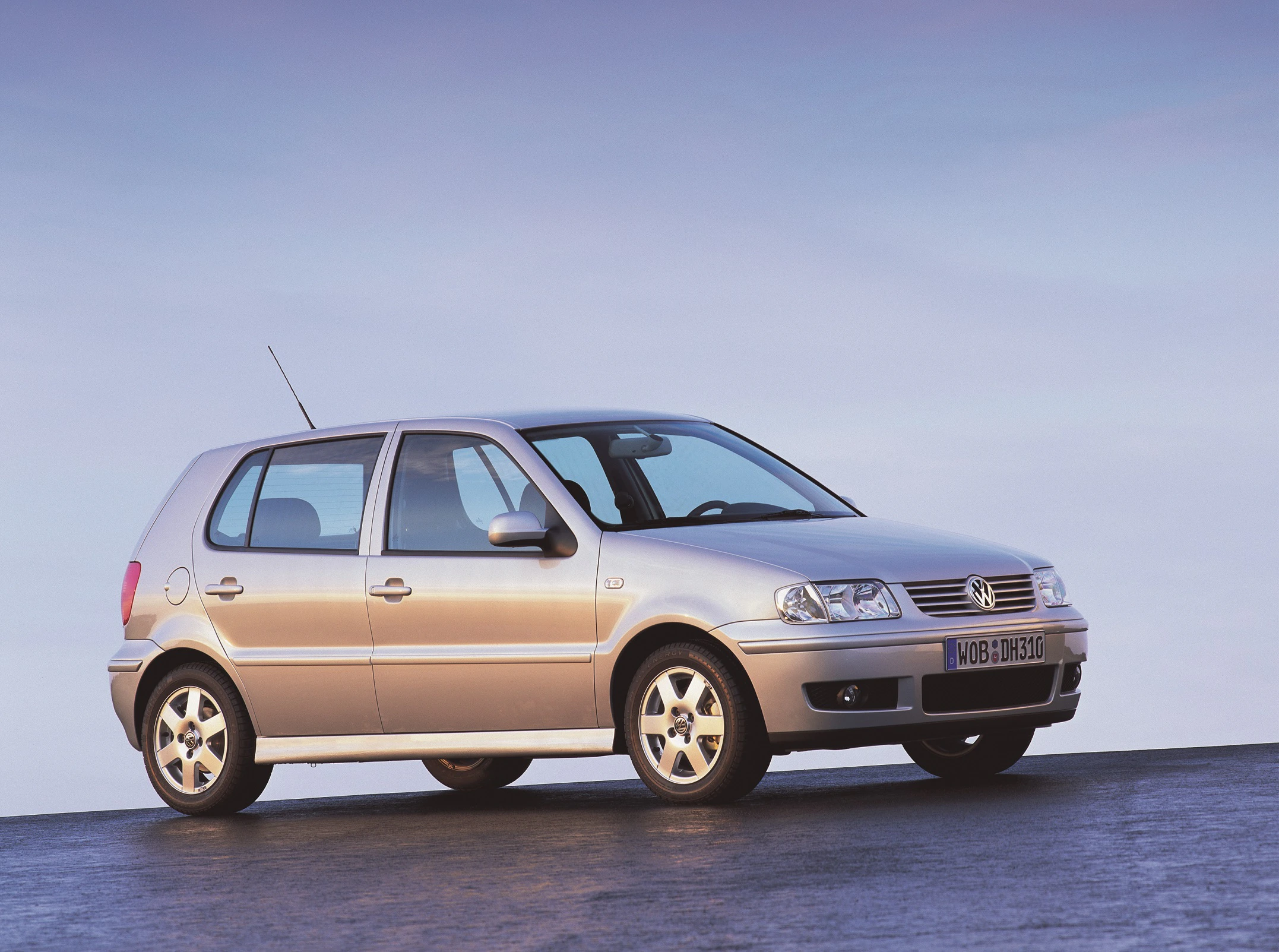 Silberner Volkswagen Polo, fünftürig, steht auf einer asphaltierten Fläche vor blauem Himmel. Hintergrund leicht bewölkt, Tageslicht.