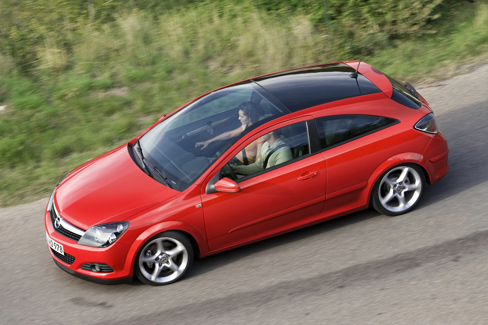 Roter Opel Astra dreitürig fährt auf einer Landstraße, Fahrer sitzt am Steuer. Im Hintergrund sind Bäume und Gras zu sehen.