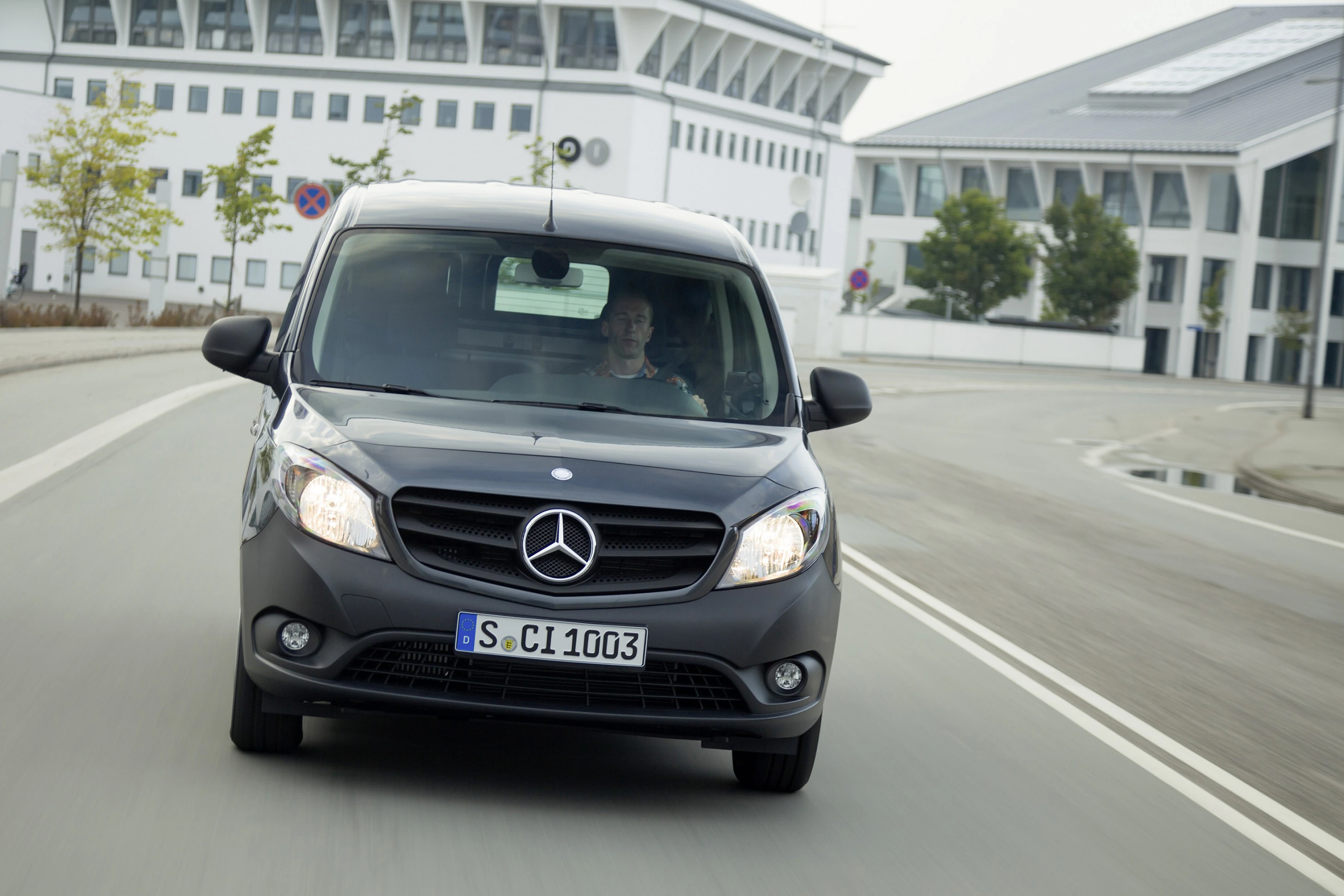 Ein schwarzer Mercedes-Benz Citan fährt auf einer urbanen Straße, moderne Gebäude im Hintergrund.