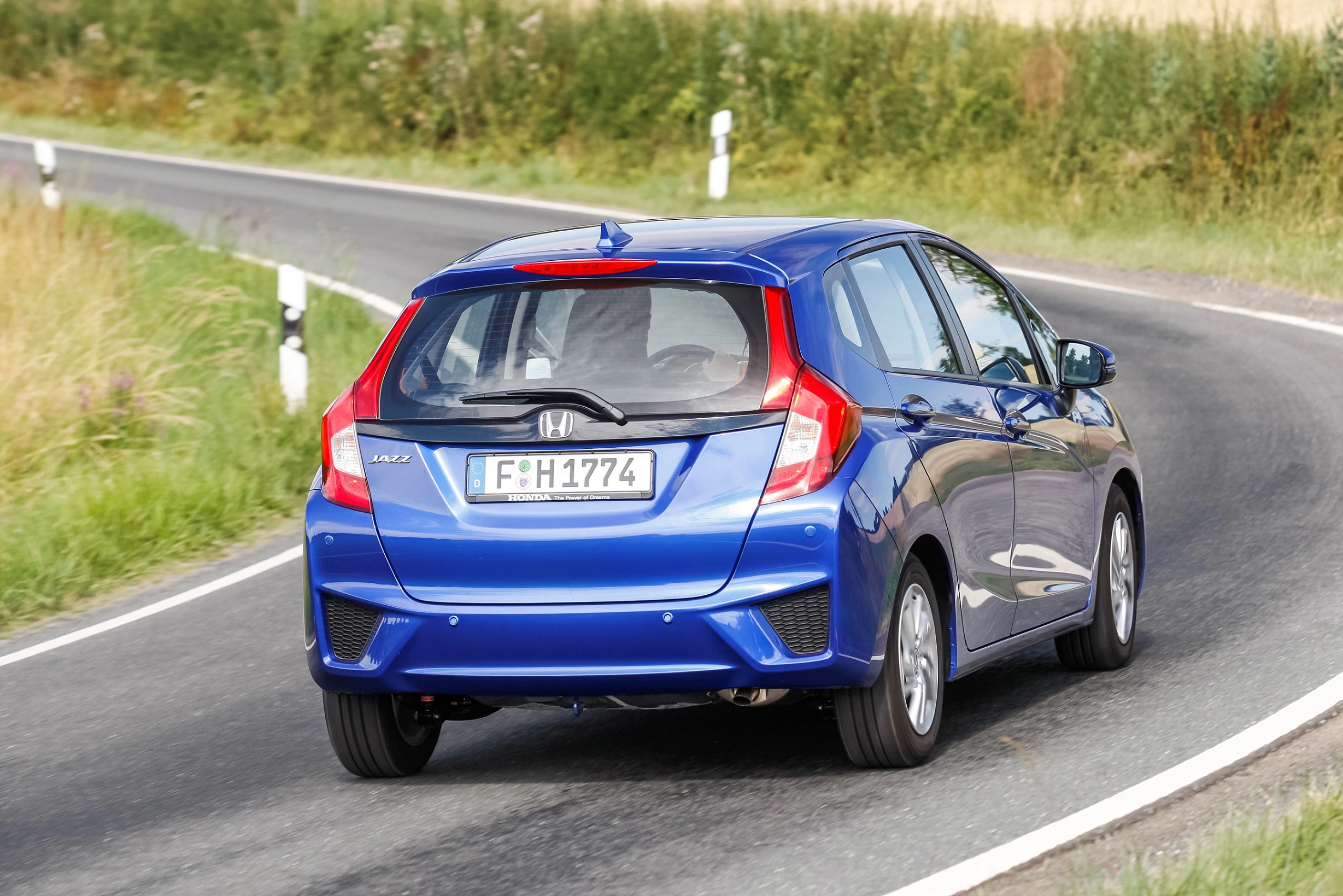 Blauer Honda Jazz fährt auf einer kurvigen Landstraße. Im Hintergrund sind Wiesen und ein heller Himmel zu sehen.