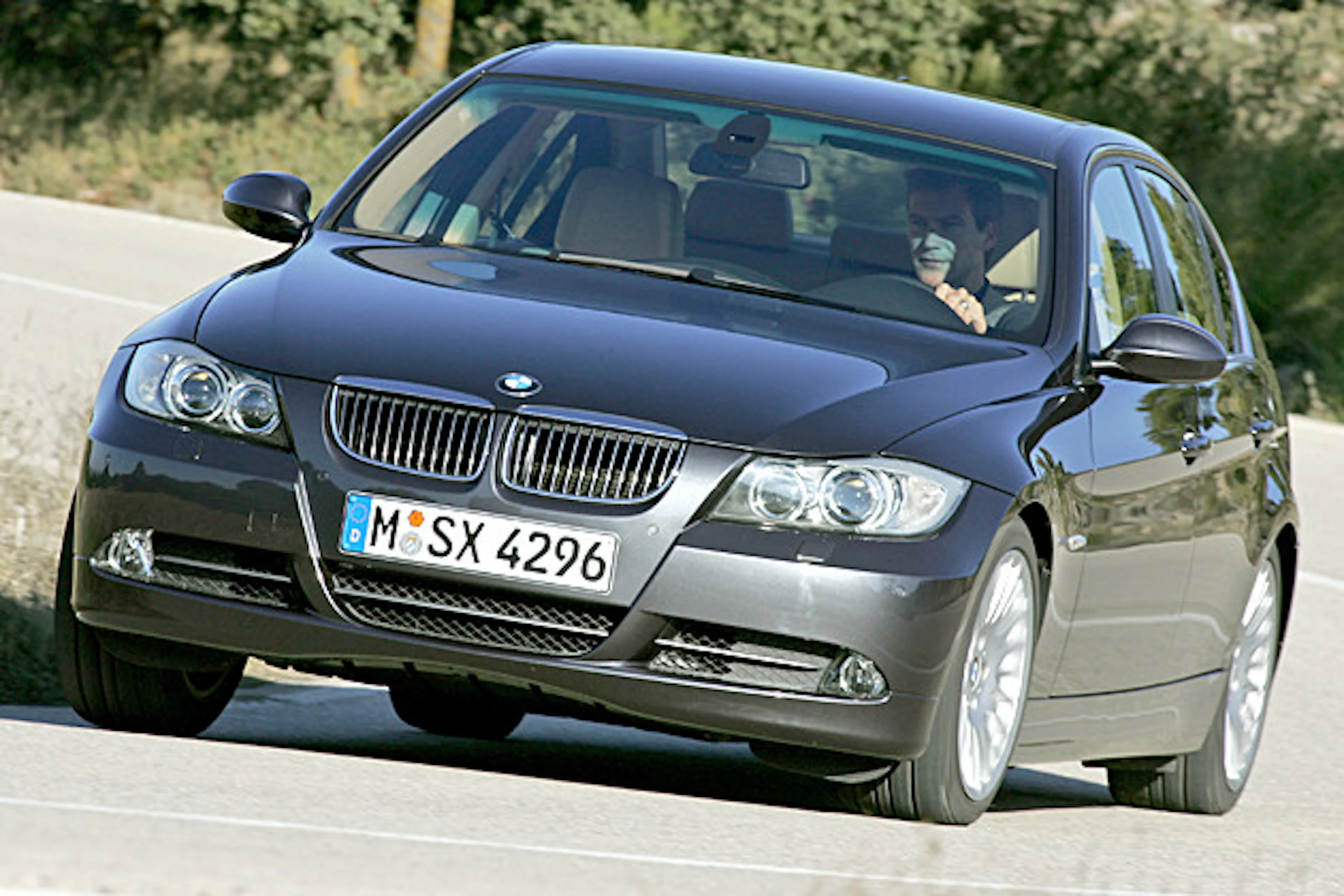 Ein schwarzer BMW fährt auf einer Landstraße. Der Fahrer ist sichtbar. Die Straße hat eine leichte Kurve und Bäume im Hintergrund.