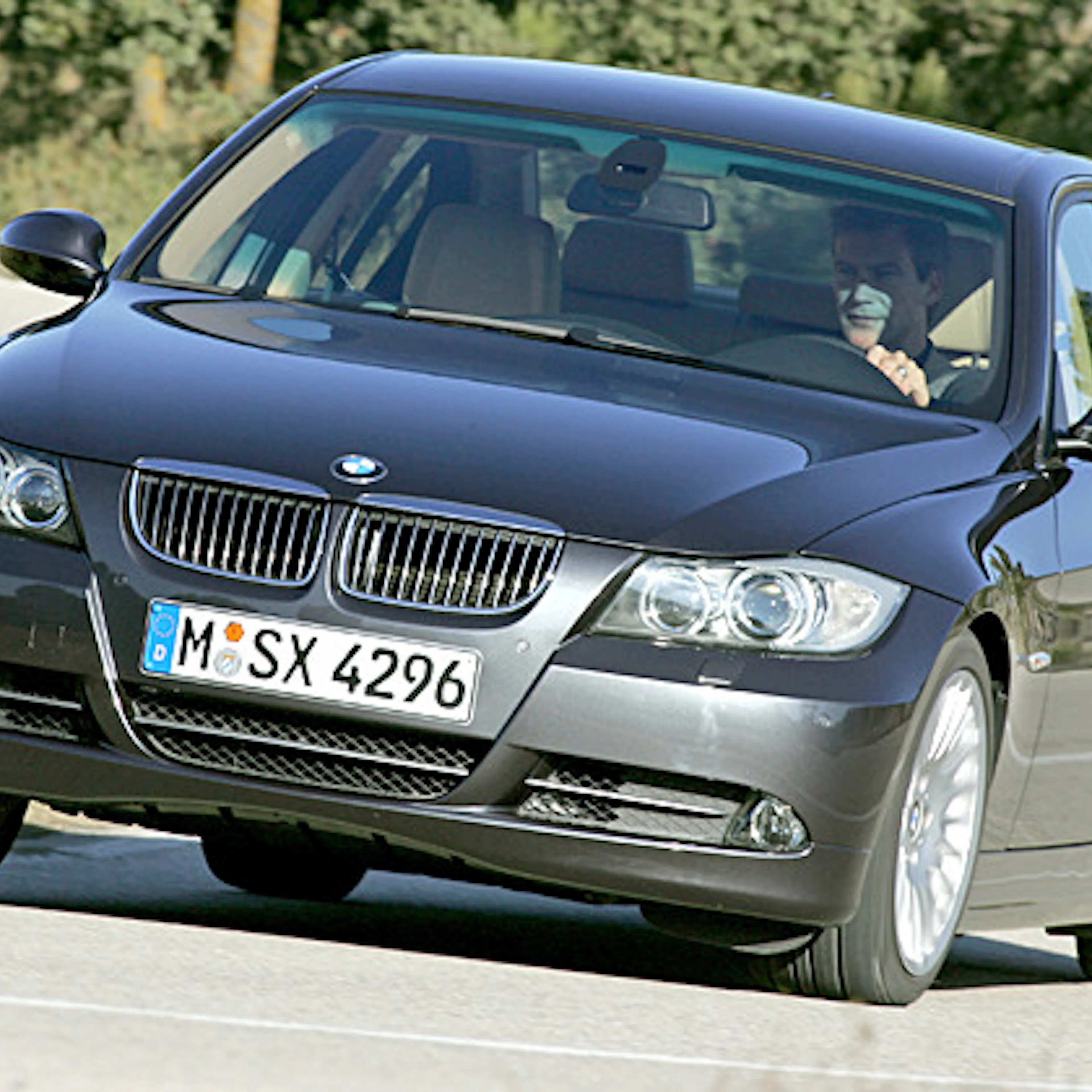 Ein schwarzer BMW fährt auf einer Landstraße. Der Fahrer ist sichtbar. Die Straße hat eine leichte Kurve und Bäume im Hintergrund.