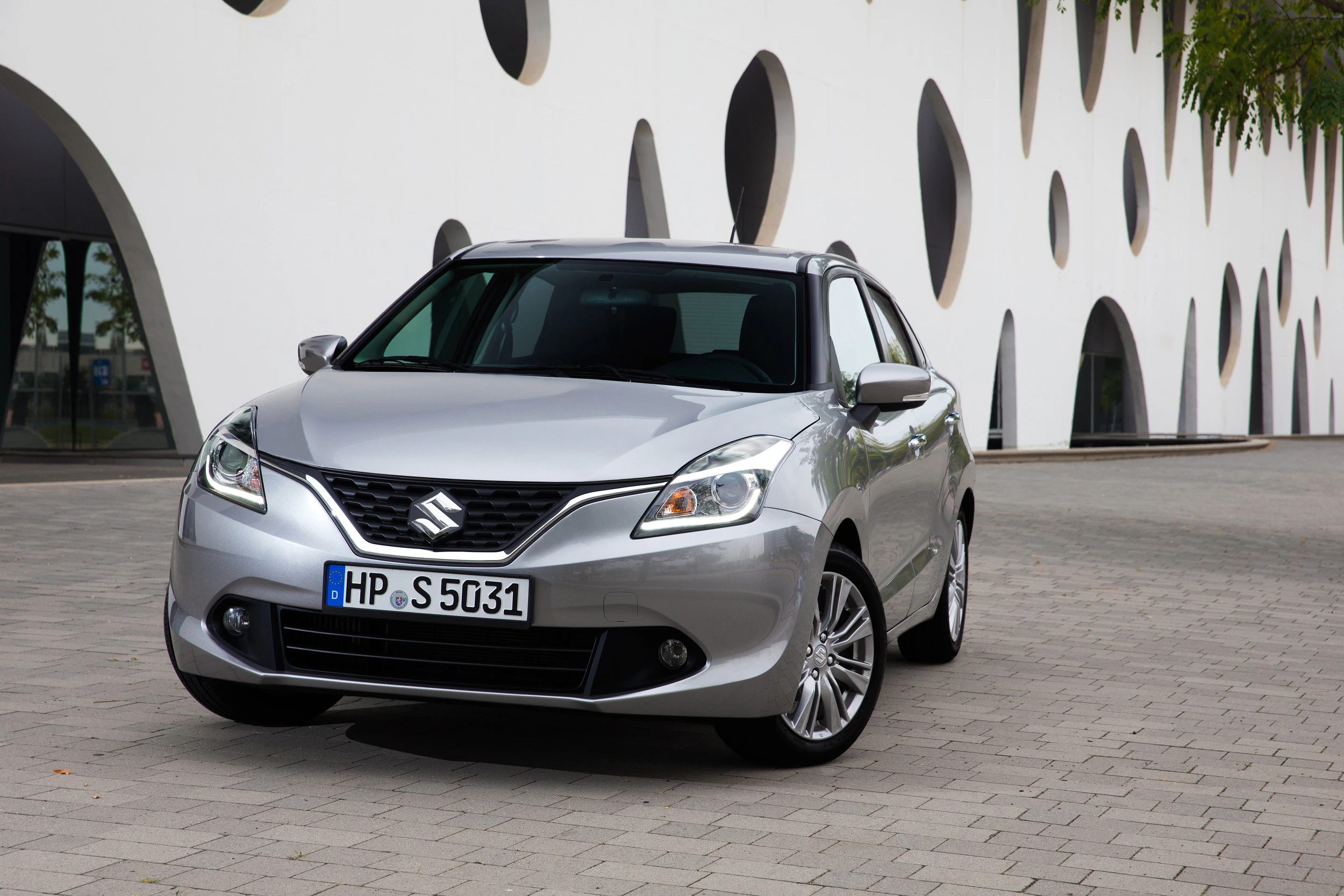 Silberner Suzuki Baleno steht auf einer gepflasterten Fläche vor einem modernen Gebäude mit runden Fenstern.