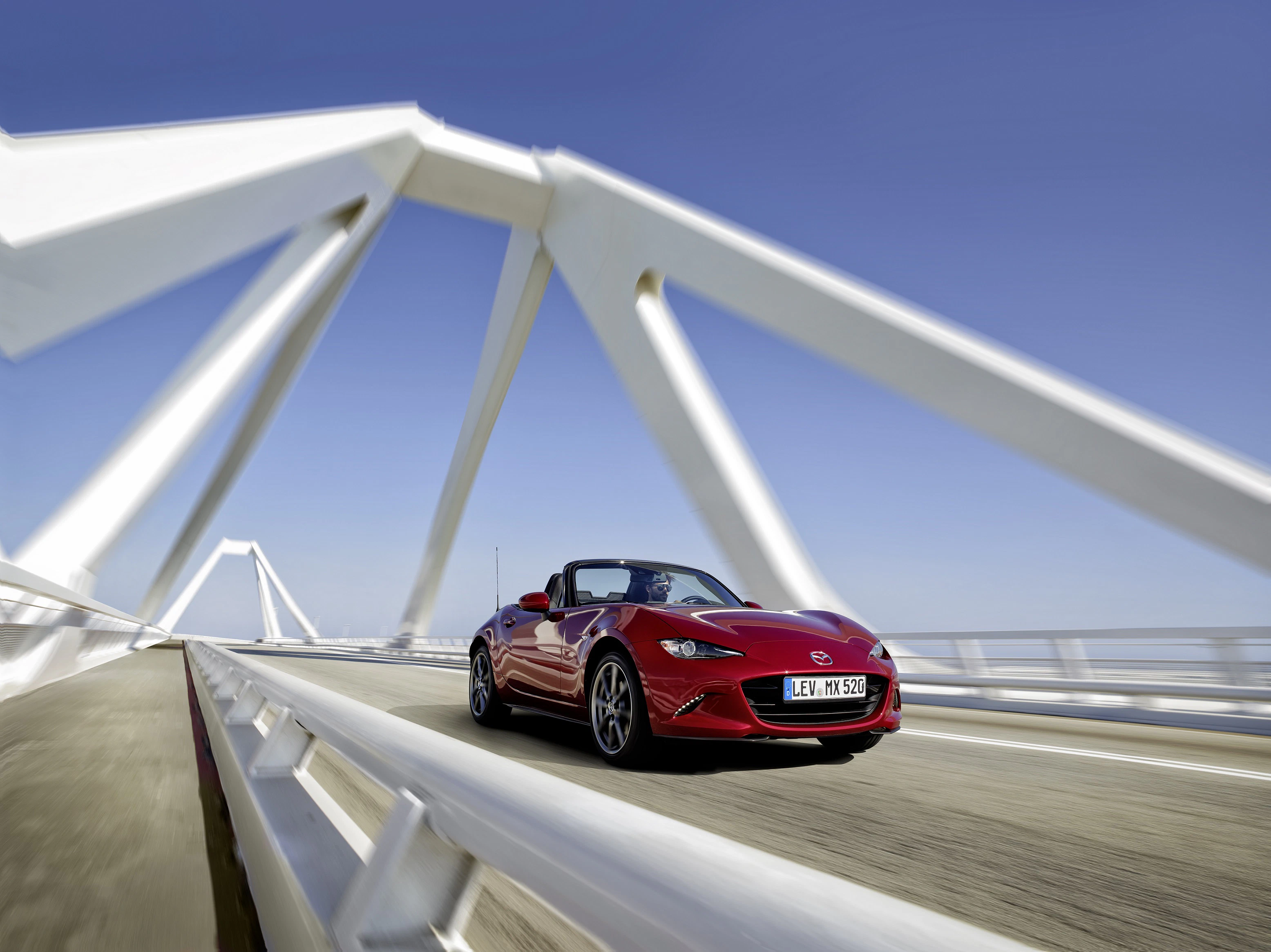 Roter Cabrio-Sportwagen fährt über moderne weiße Brücke bei blauem Himmel.