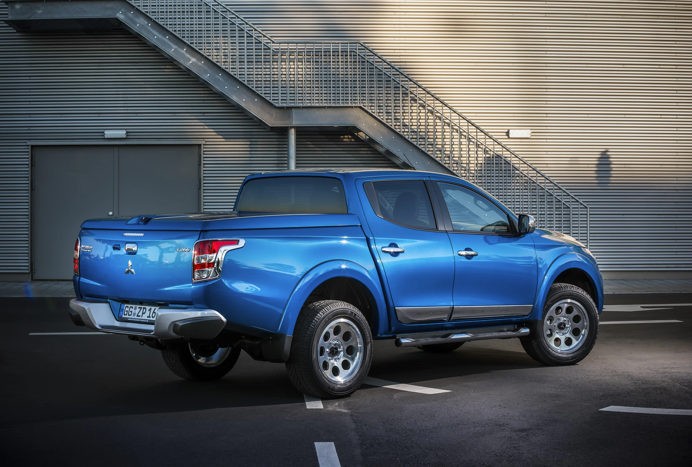 Blauer Pickup-Truck steht auf einem Parkplatz vor einem modernen Gebäude mit Metalltreppe und grauen Wänden.