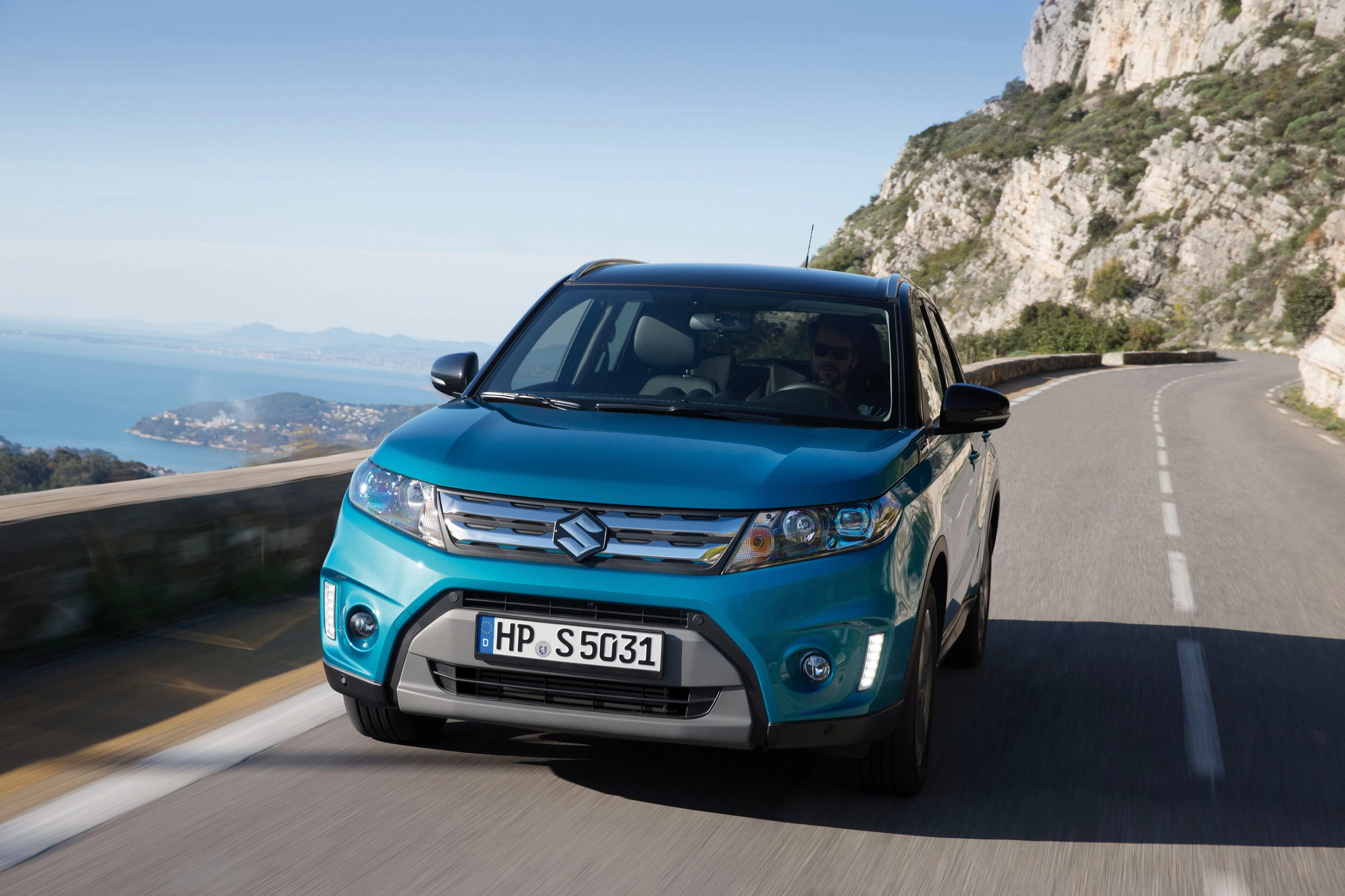 Blauer Suzuki SUV fährt auf einer kurvenreichen Küstenstraße entlang einer Felswand mit Meerblick im Hintergrund.