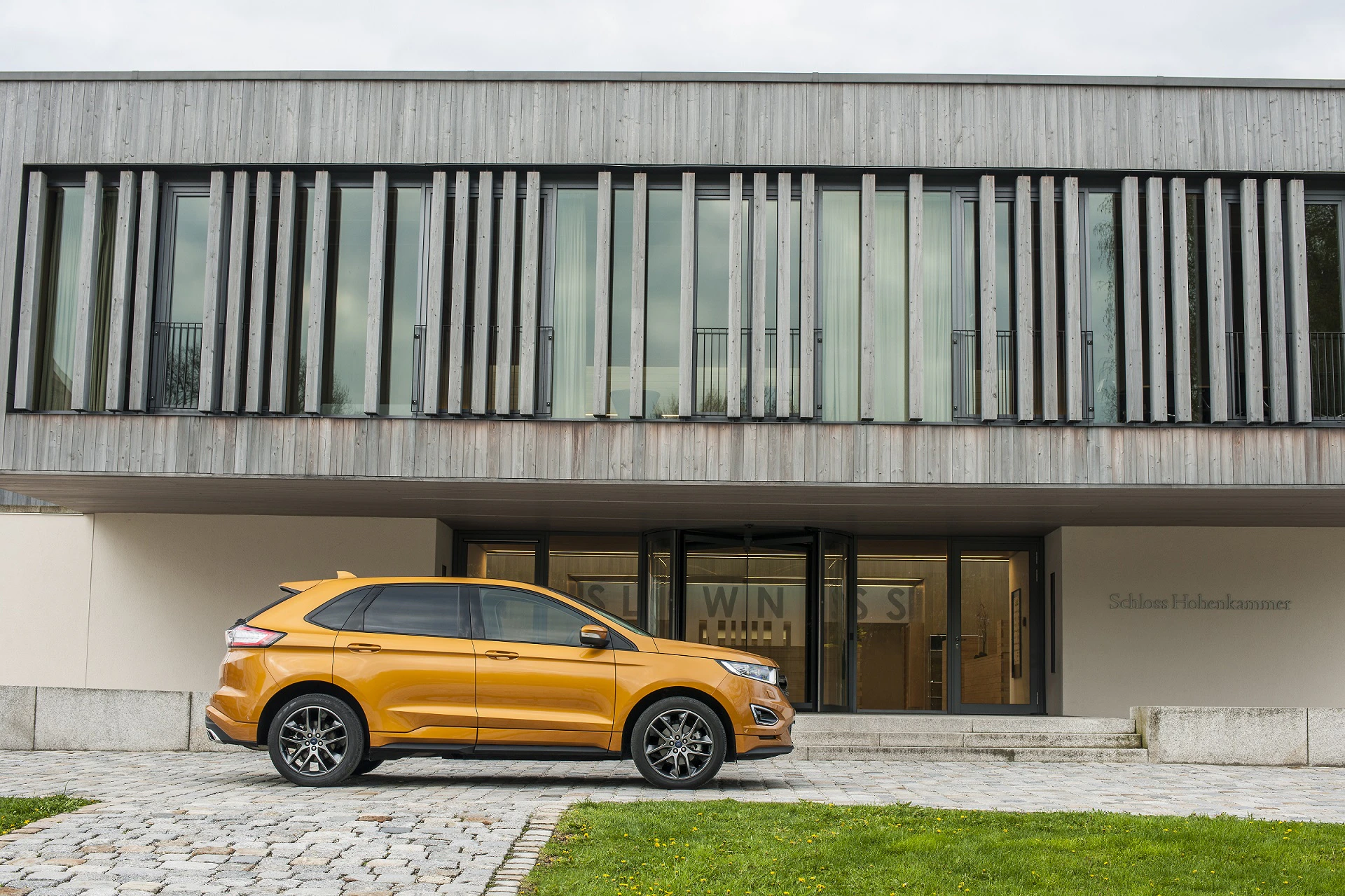 Ein goldener SUV parkt vor einem modernen, zweistöckigen Gebäude mit großen Fenstern und vertikalen Holzelementen.