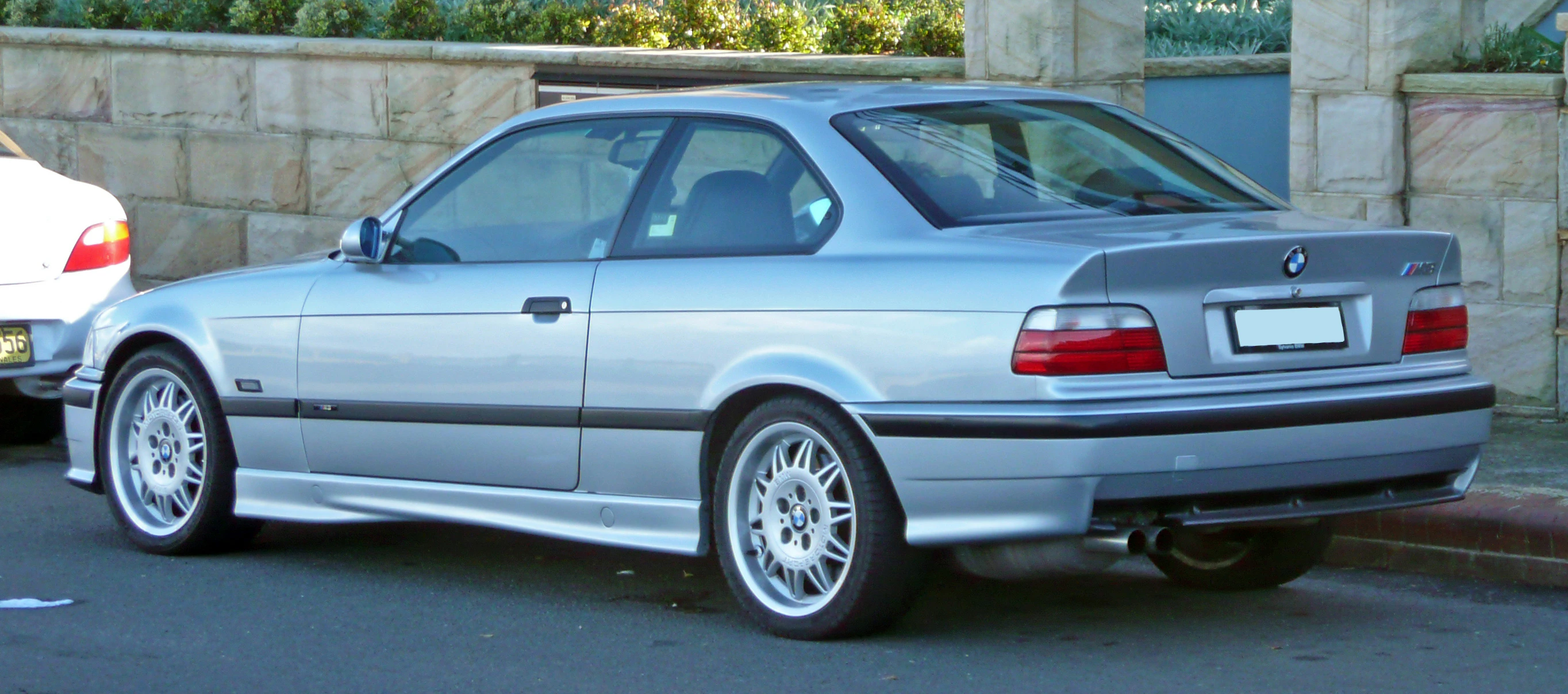 Silberner BMW E36 Coupe, geparkt auf der Straße. Er hat schwarze Zierleisten, Leichtmetallräder und getönte Fenster.