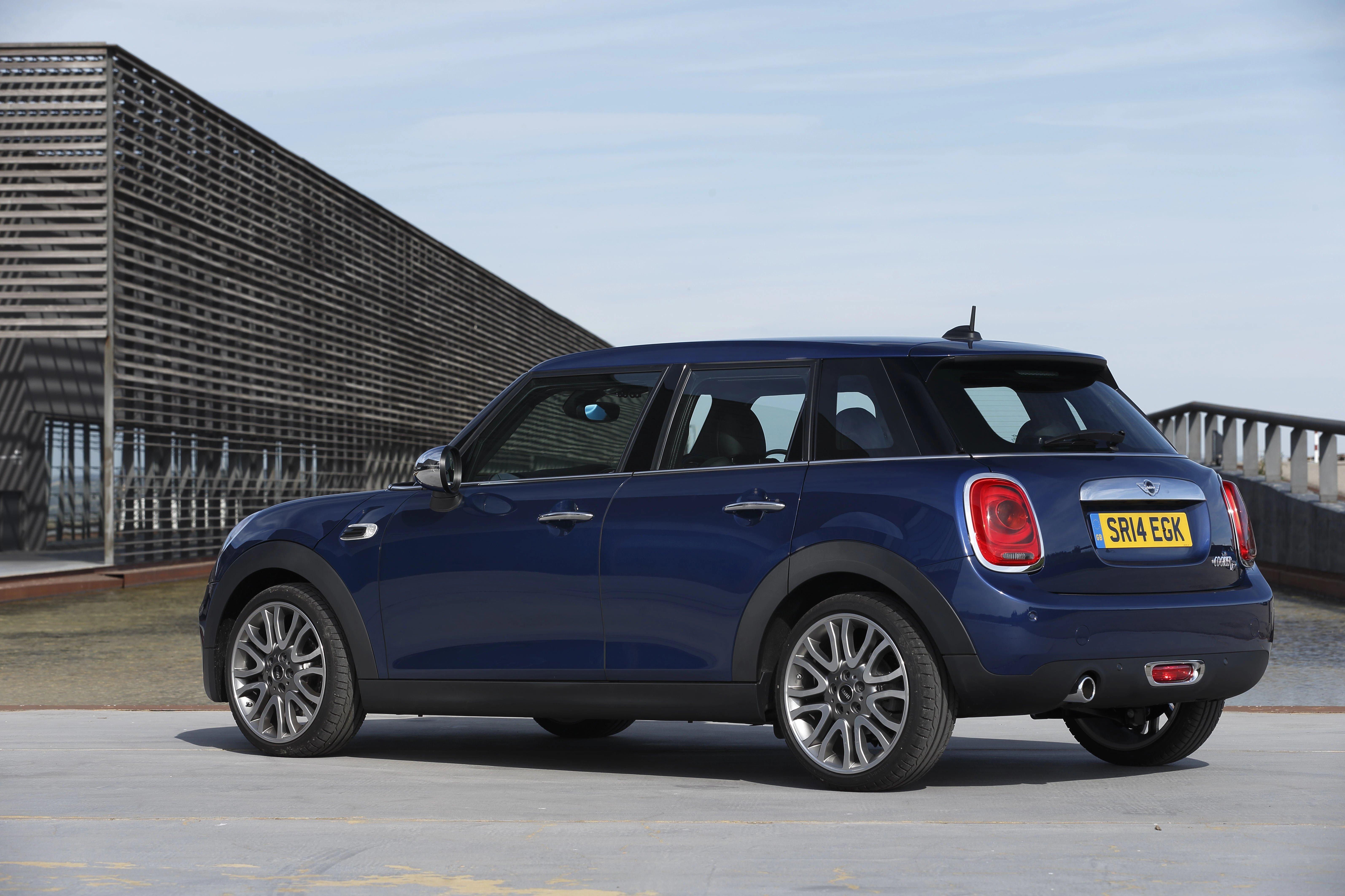 Blauer Mini Cooper Clubman vor modernem Gebäude, seitlich von hinten fotografiert, auf einer Betonfläche mit klarem Himmel im Hintergrund.