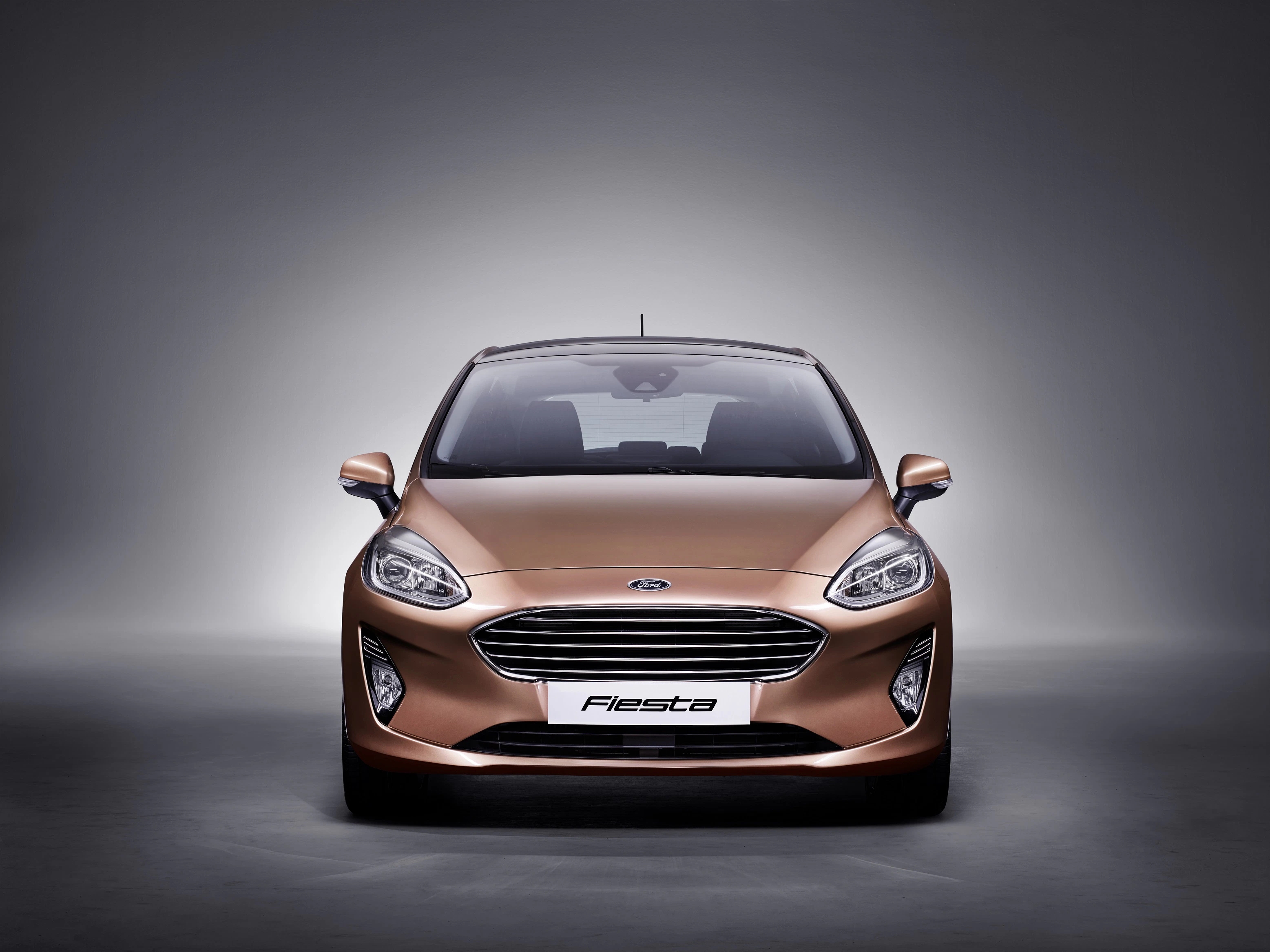 Frontansicht eines bronzefarbenen Ford Fiesta vor grauem Hintergrund. Moderne Scheinwerfer und markanter Kühlergrill heben sich hervor.