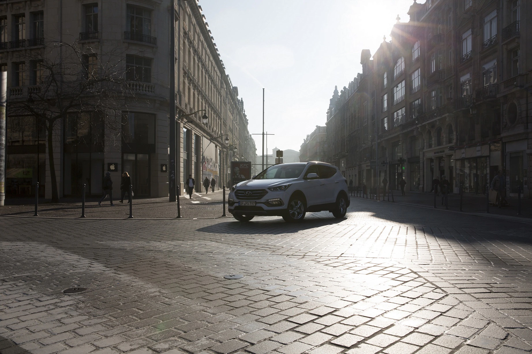 Weißes Auto fährt bei Sonnenuntergang durch eine Kopfsteinpflasterstraße, umgeben von historischen Gebäuden. Passanten im Hintergrund.