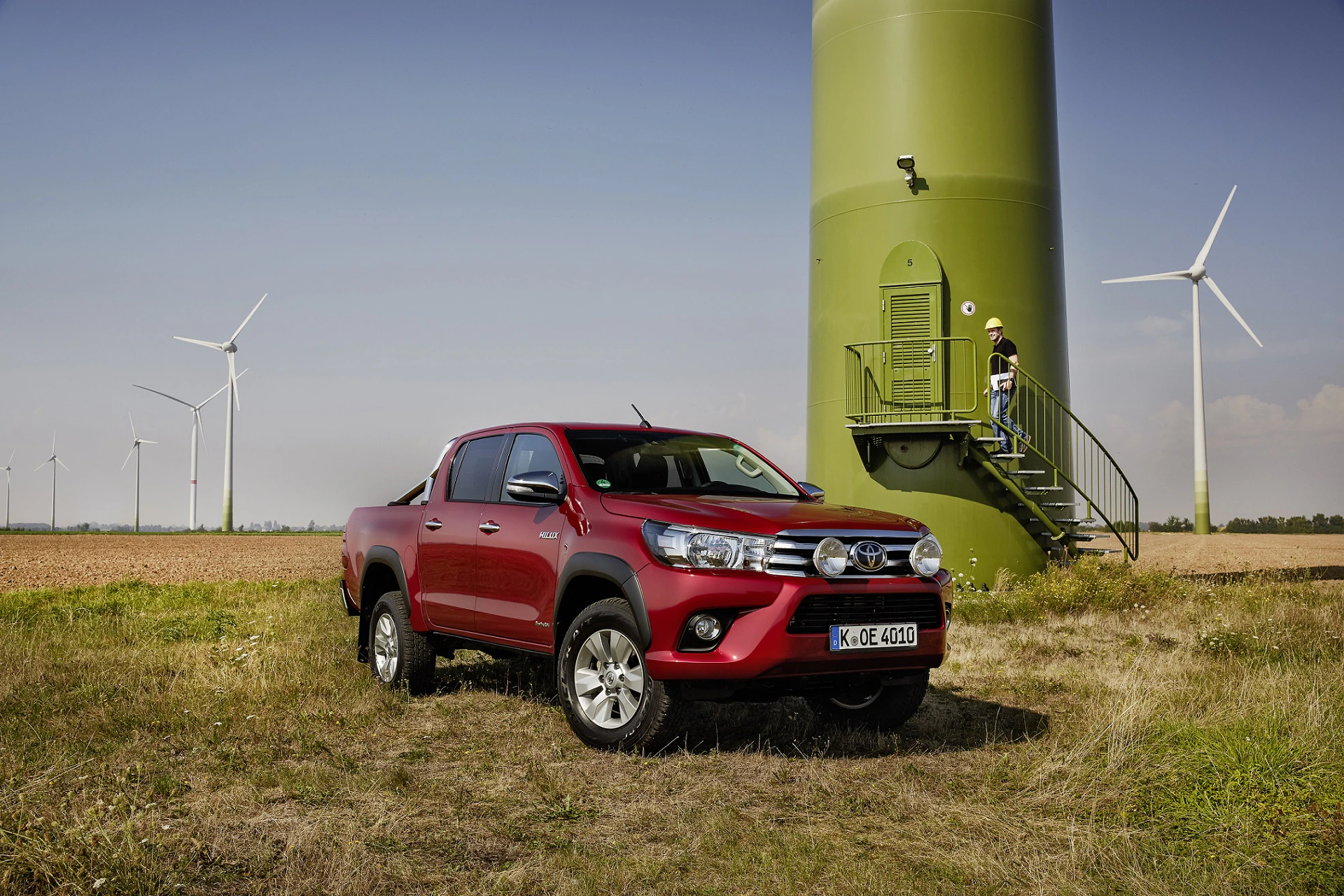 Roter Pickup-Truck parkt auf einem Feld vor einem grünen Windrad. Frau steigt eine Treppe hinauf. Mehrere Windräder im Hintergrund.