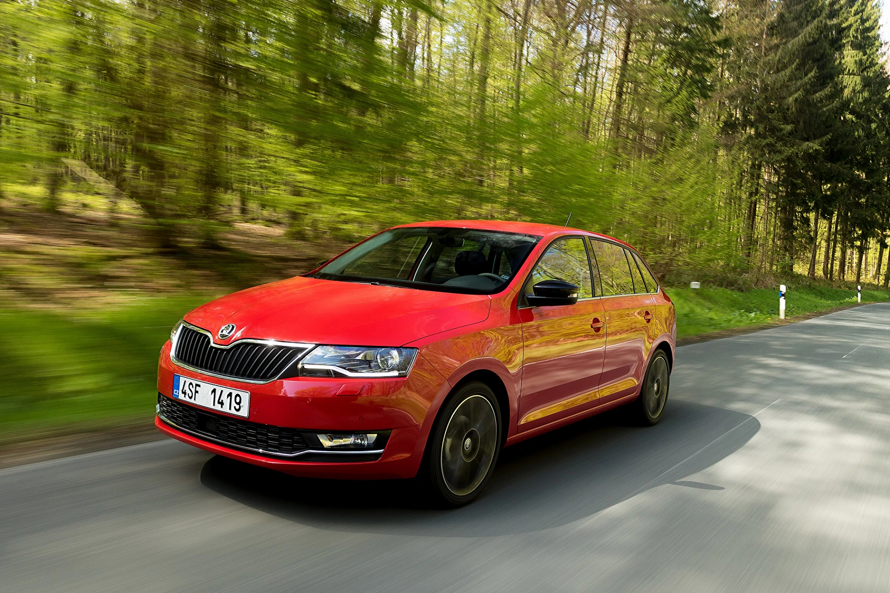Roter Škoda Rapid fährt auf einer Landstraße durch einen grünen Wald, mit verschwommenem Hintergrund, der Dynamik zeigt.