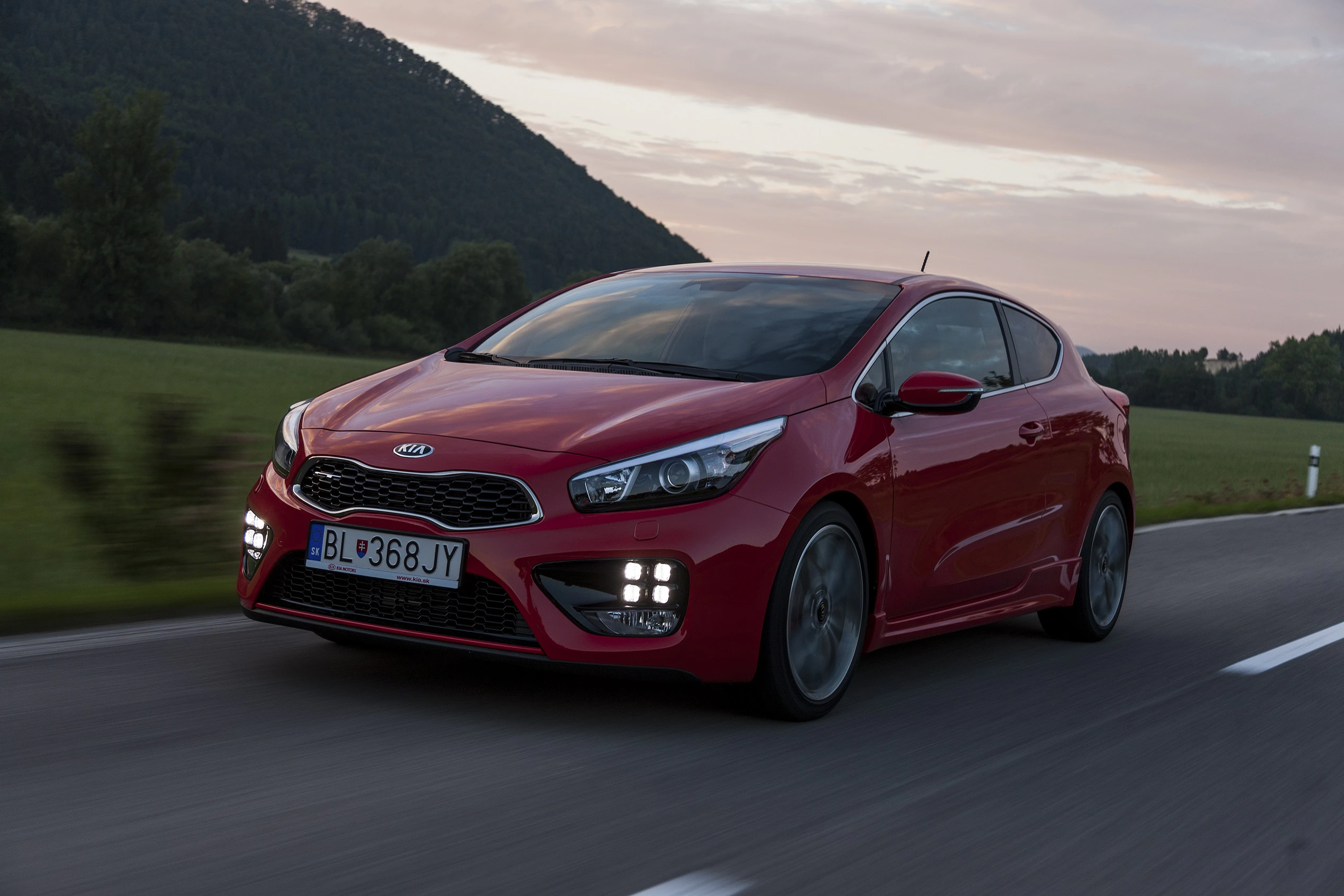 Roter Kia Cee'd fährt auf einer Landstraße bei Sonnenuntergang; umgeben von grüner Landschaft und Hügeln.