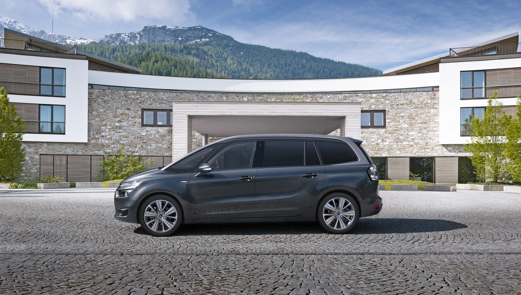 Schwarzer Minivan steht auf Kopfsteinpflaster vor modernem Gebäude mit Bergkulisse und blauen Himmel im Hintergrund.