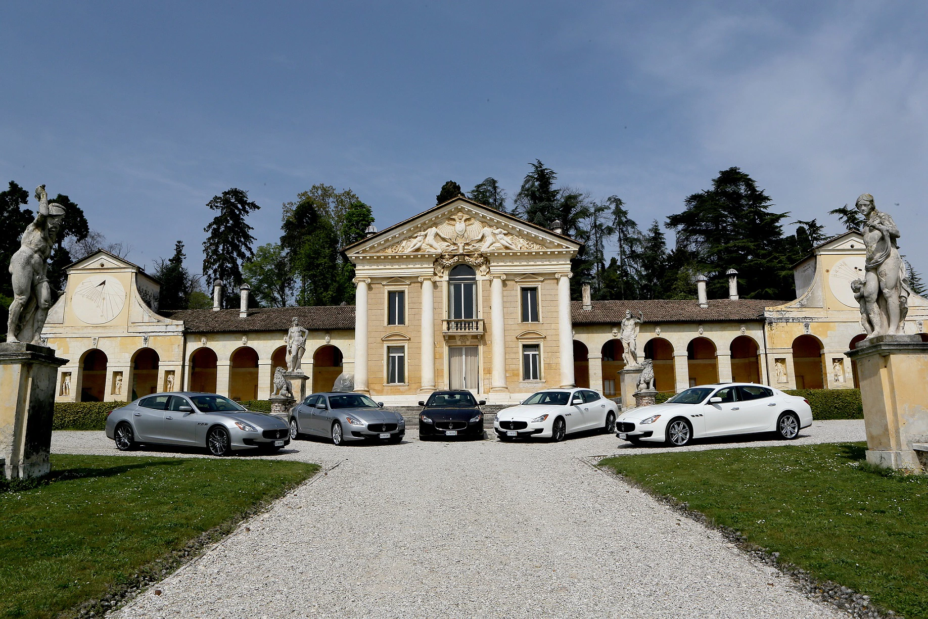 Fünf Luxusautos vor einer historischen Villa mit symmetrischen Statuen und einem Kiesweg, umgeben von Bäumen und blauem Himmel.