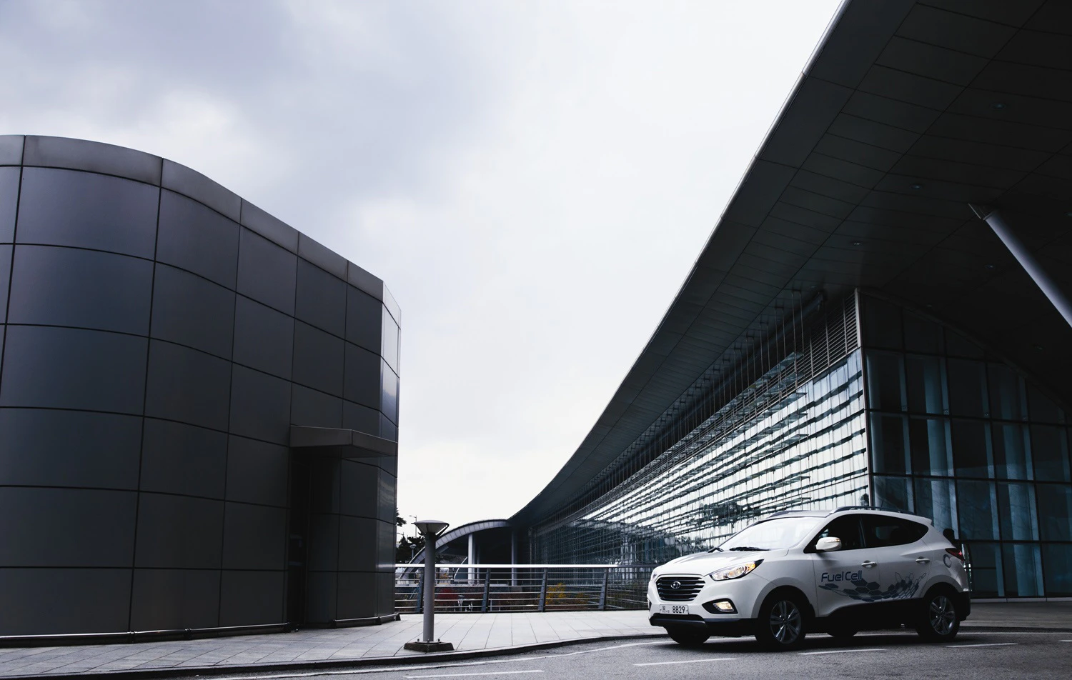 Ein weißes Auto fährt an moderner Architektur mit geschwungenen Glasfassaden vorbei; bewölkter Himmel im Hintergrund.