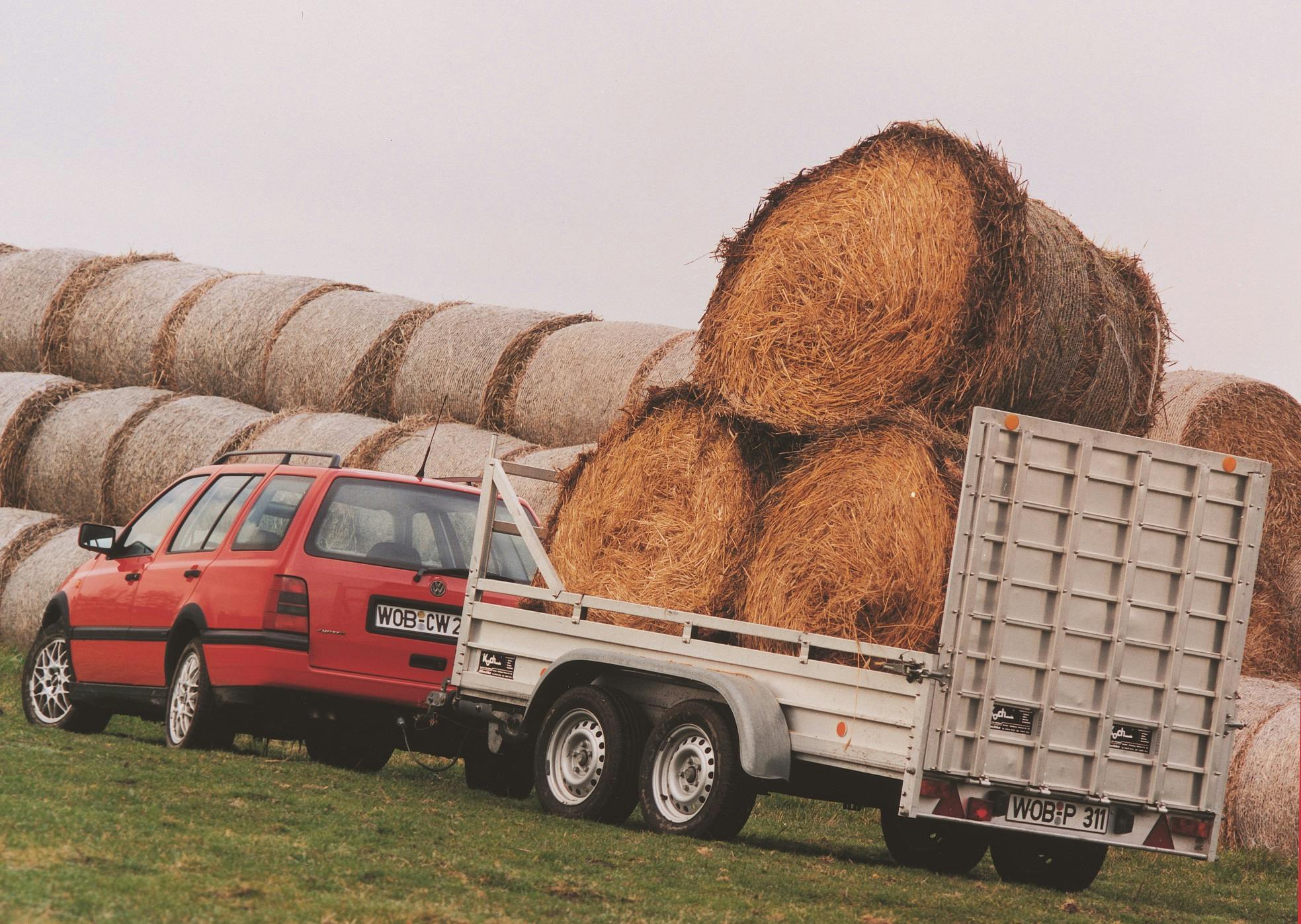 Roter Kombi mit Anhänger transportiert große Strohballen. Im Hintergrund liegen weitere Strohballen auf einem Feld.