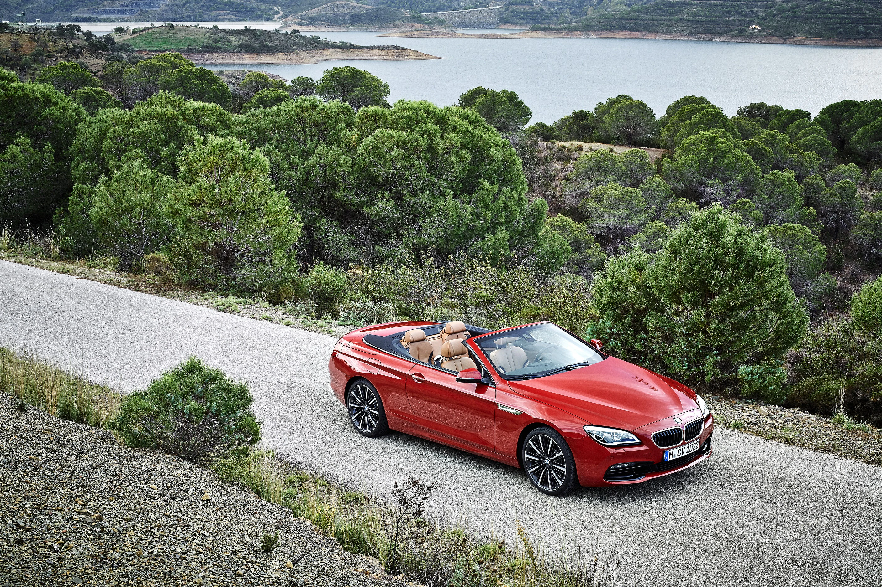 Rotes Cabrio auf einer Landstraße, umgeben von Bäumen, mit einem See im Hintergrund. Fahrer genießt die Fahrt bei sonnigem Wetter.