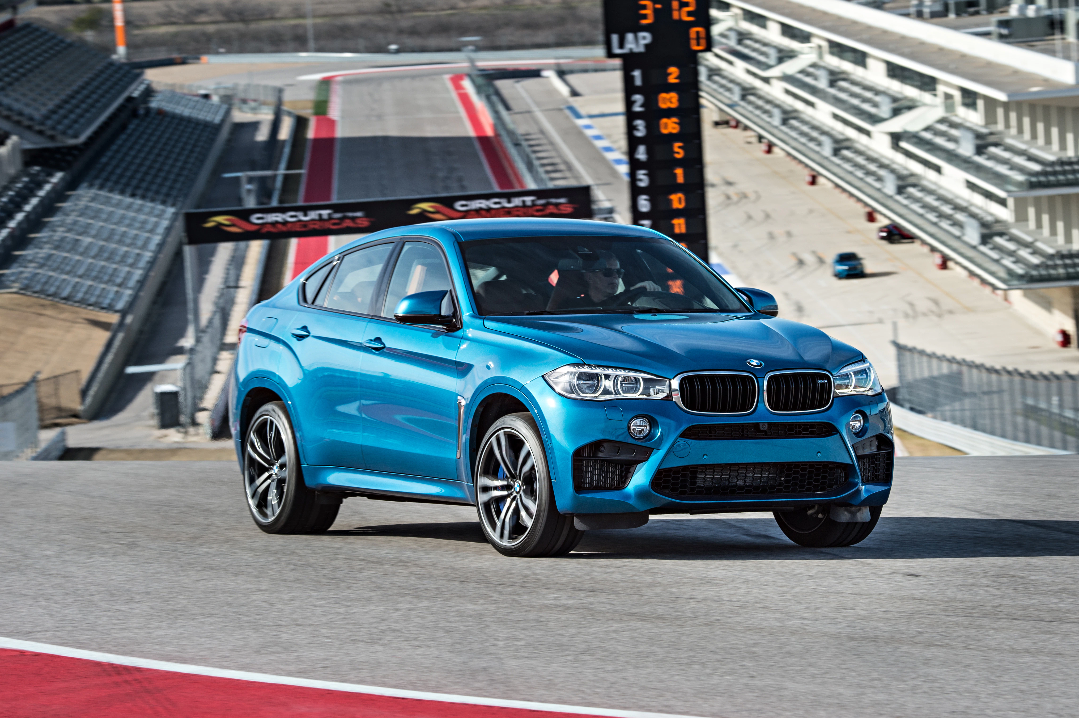 Blauer BMW X6 M fährt auf einer Rennstrecke. Im Hintergrund sind Tribünen und Anzeigetafel zu sehen. Sportliche Atmosphäre.