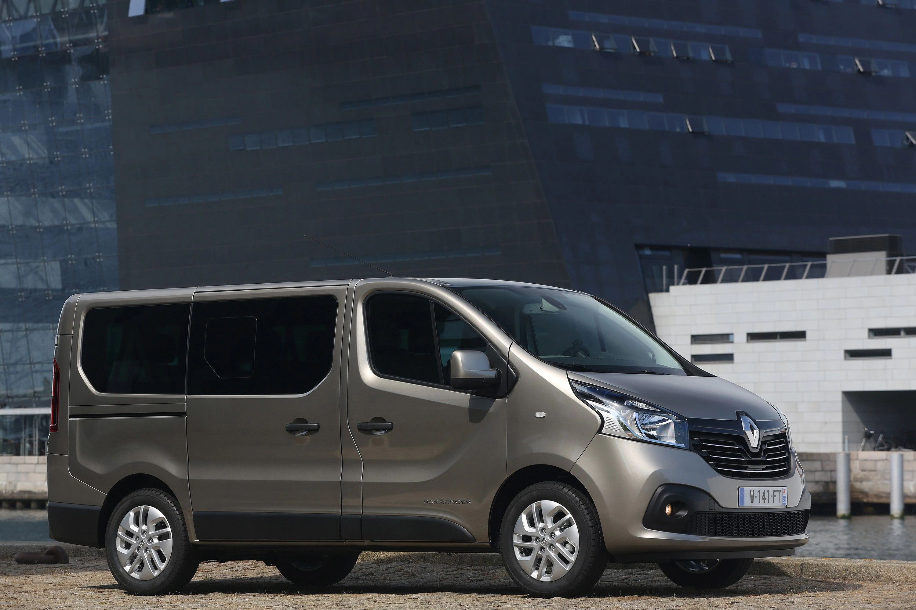 Grauer Renault-Van parkt vor modernem, schwarzem Gebäude und Wasser. Fahrzeug seitlich von links zu sehen, urbaner Hintergrund.