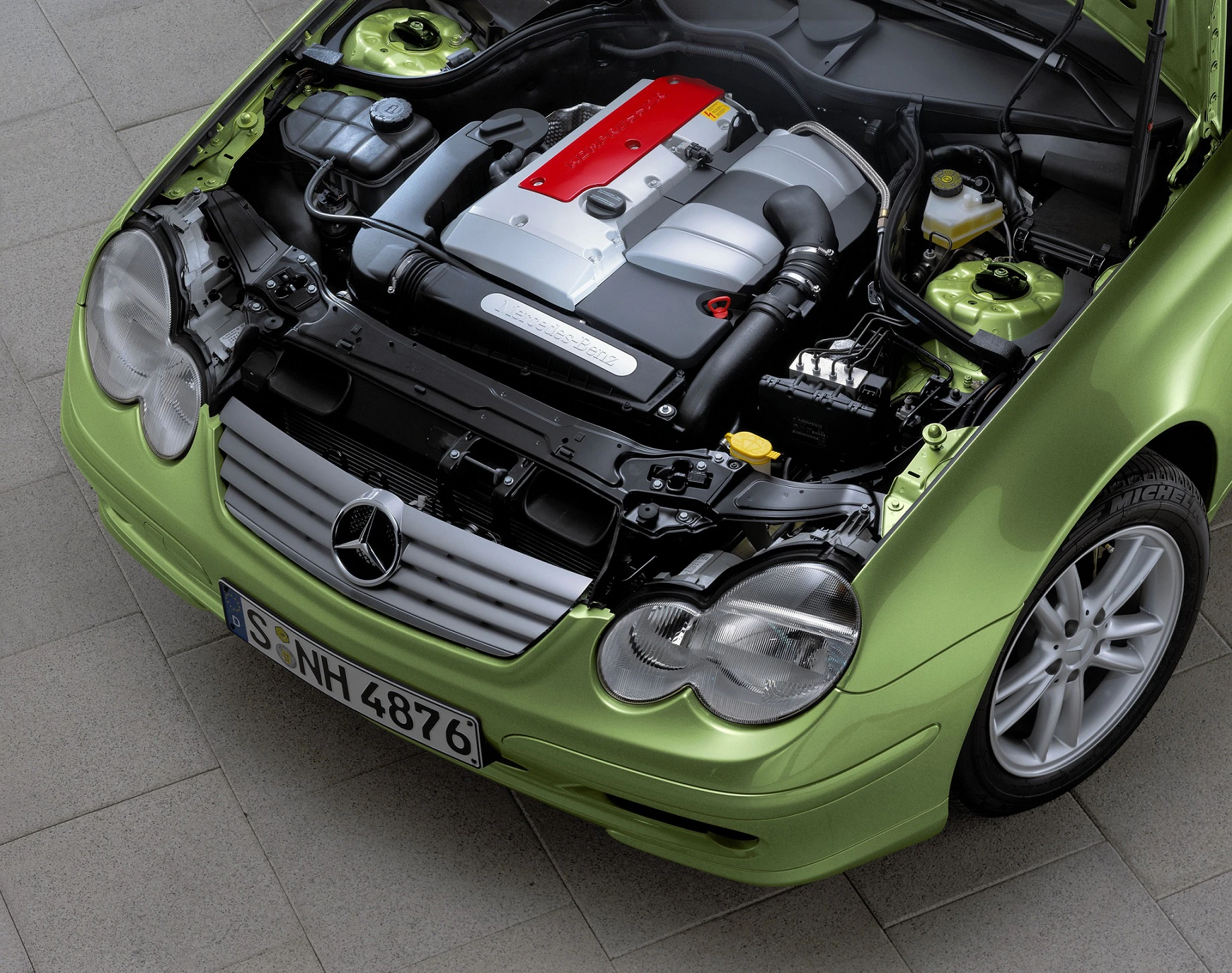 Grüner Mercedes mit geöffneter Motorhaube, zeigt den Motor und silberne Details, auf grauem Boden fotografiert.