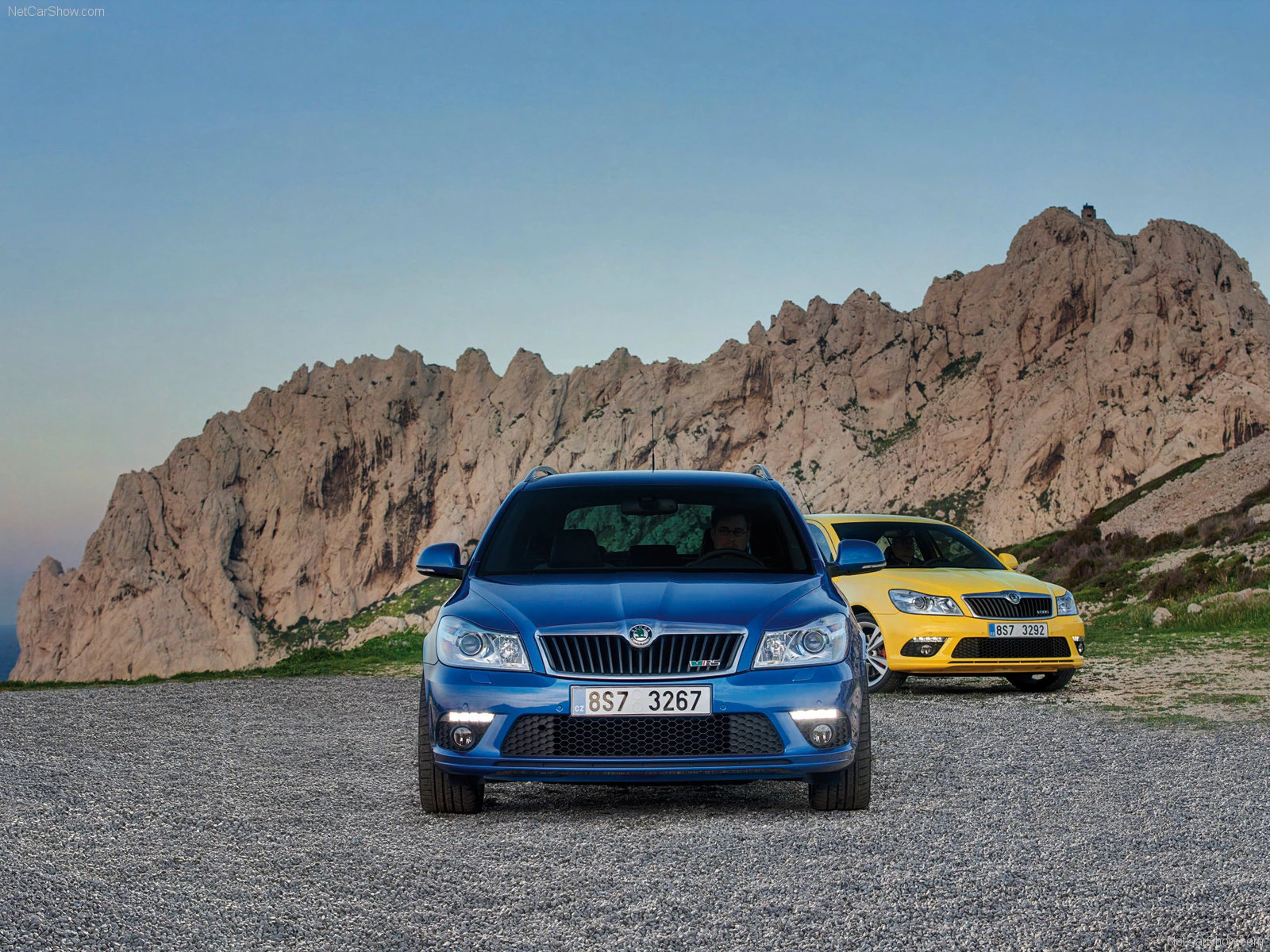 Zwei Autos, ein blaues und ein gelbes, stehen auf einem steinigen Weg vor einer spektakulären Felslandschaft unter klarem Himmel.