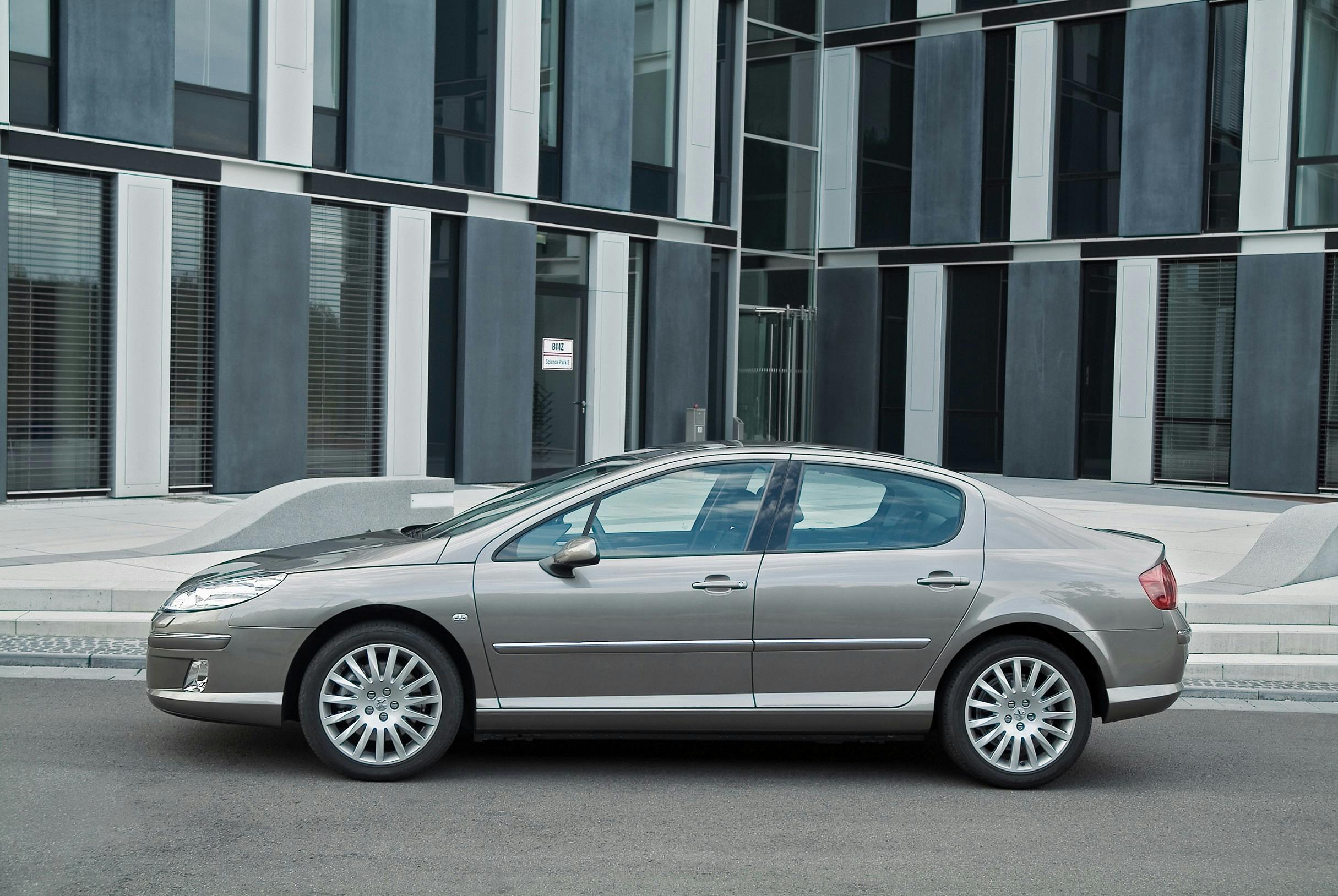 Graues Auto parkt vor einem modernen Gebäude mit großen Fenstern und vertikalen Fassadenelementen auf Asphaltstraße.