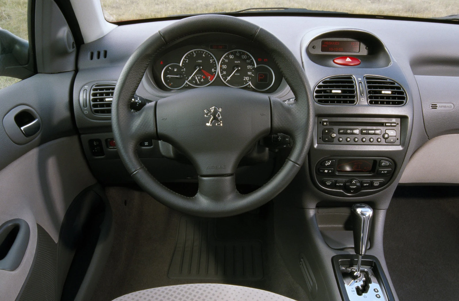 Innenraum eines Autos mit Peugeot-Logo auf dem Lenkrad, Armaturenbrett mit Tacho, Radiosteuerung und Schalthebel sichtbar.