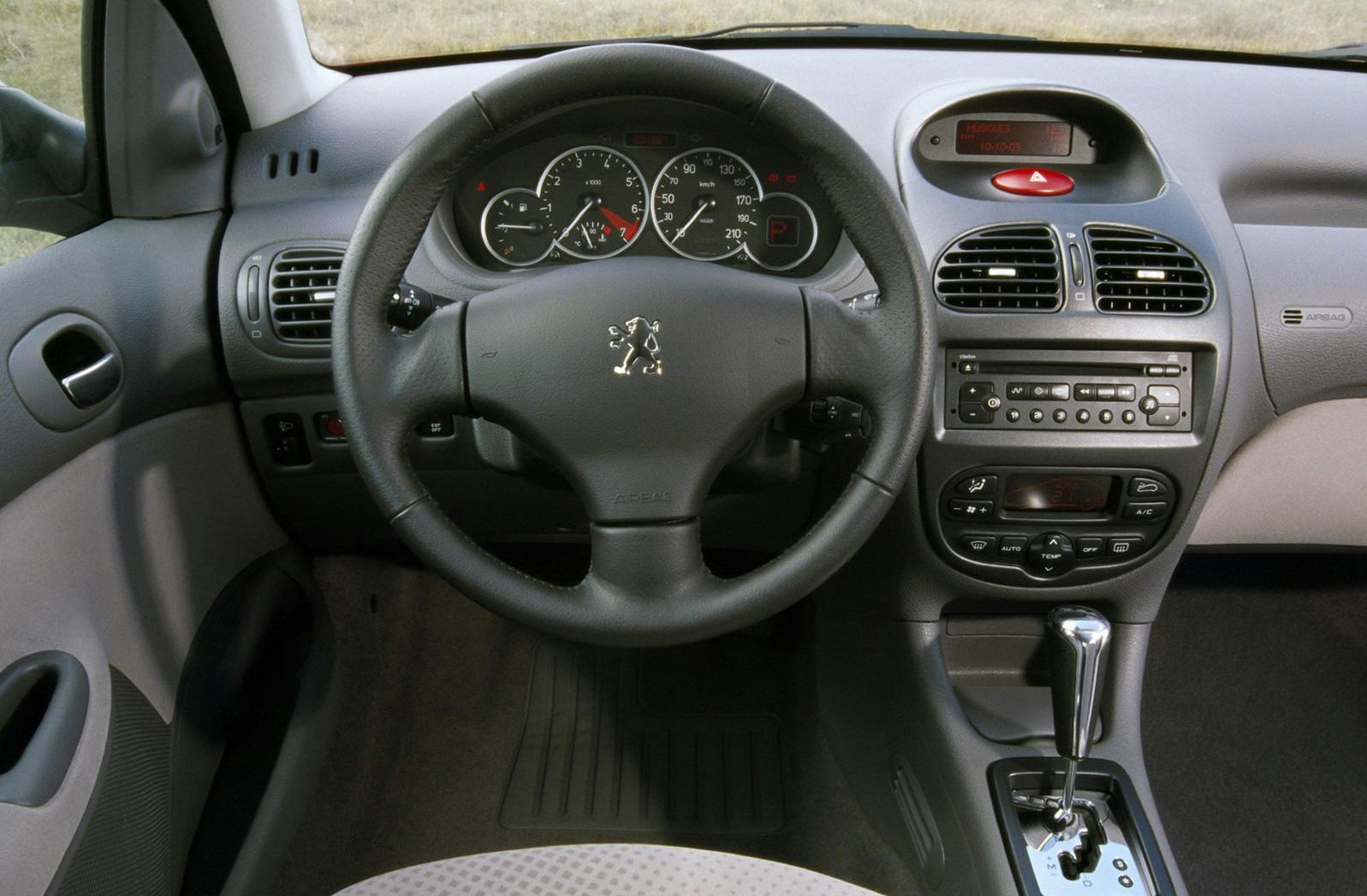 Innenraum eines Autos mit Peugeot-Logo auf dem Lenkrad, Armaturenbrett mit Tacho, Radiosteuerung und Schalthebel sichtbar.