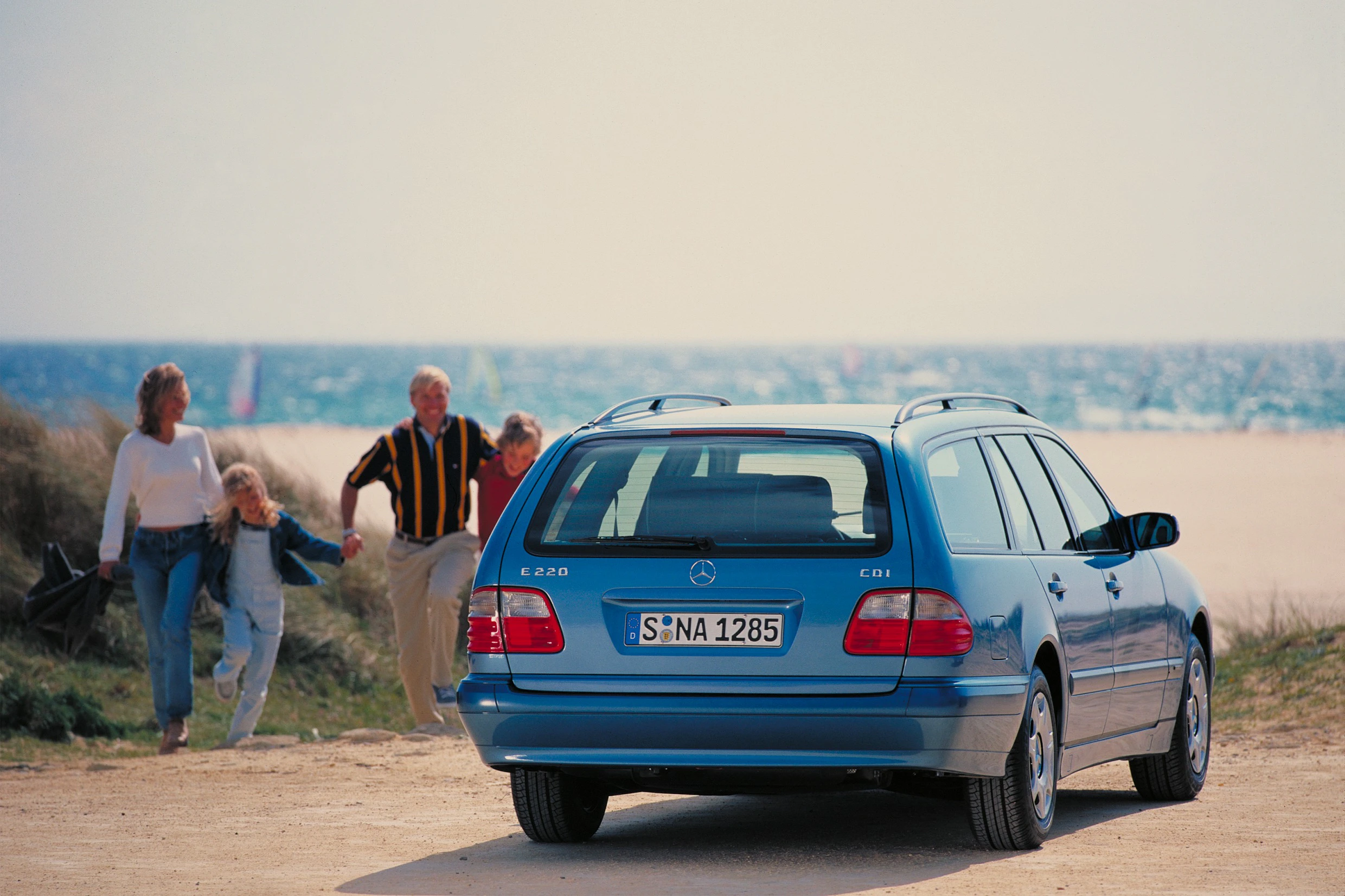 Blauer Mercedes E 220 CDI Kombi am Strand, im Hintergrund eine Familie beim Spazierengehen. Ozean und Segelboote sind sichtbar.