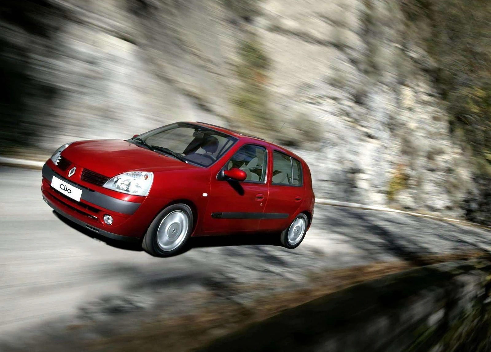 Roter Renault Clio fährt schnell auf kurviger Bergstraße, umgeben von Felsen und Bäumen.