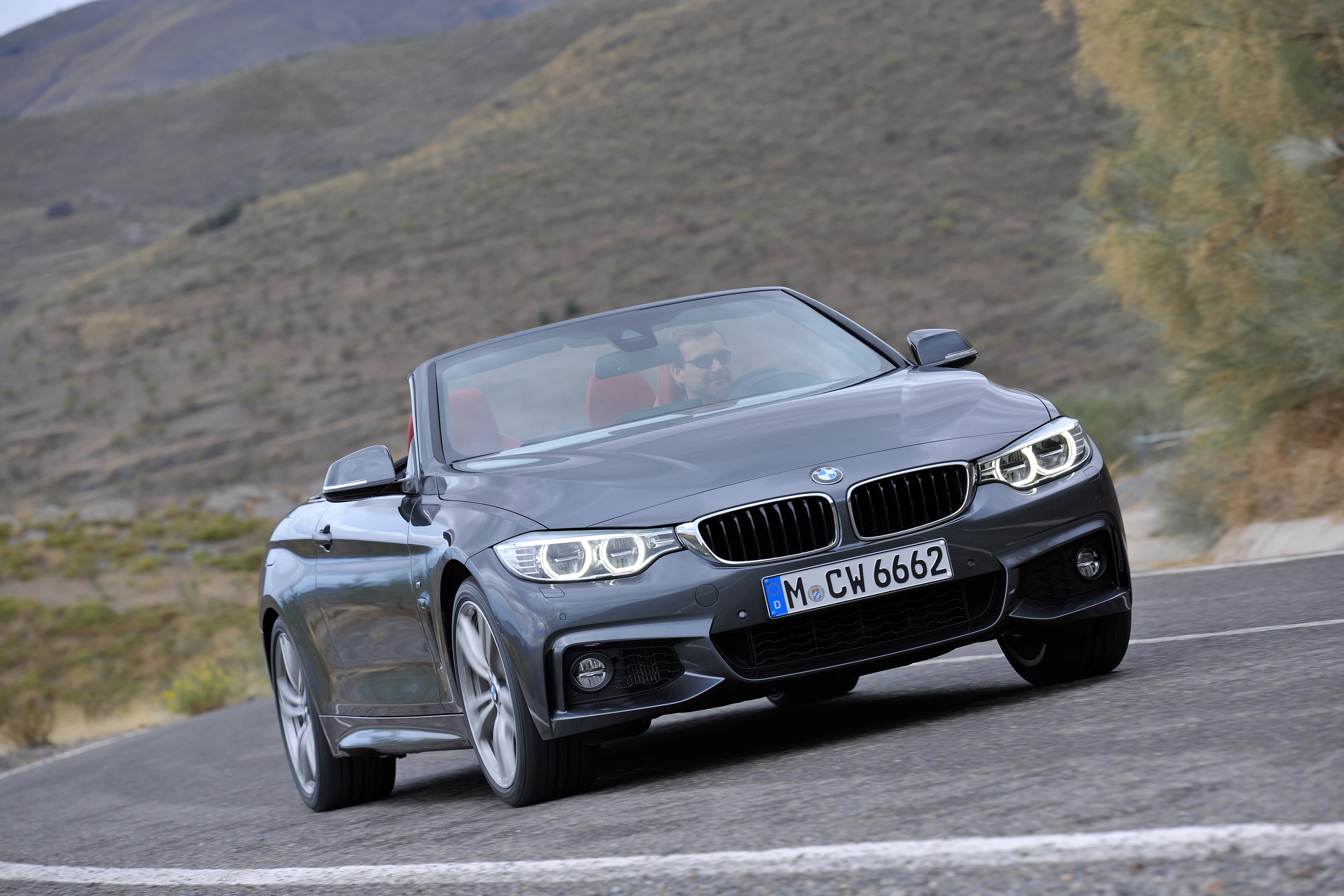 Graues BMW-Cabrio fährt auf kurviger Straße in einer bergigen Landschaft. Fahrzeugfront und Felgen sind sichtbar.