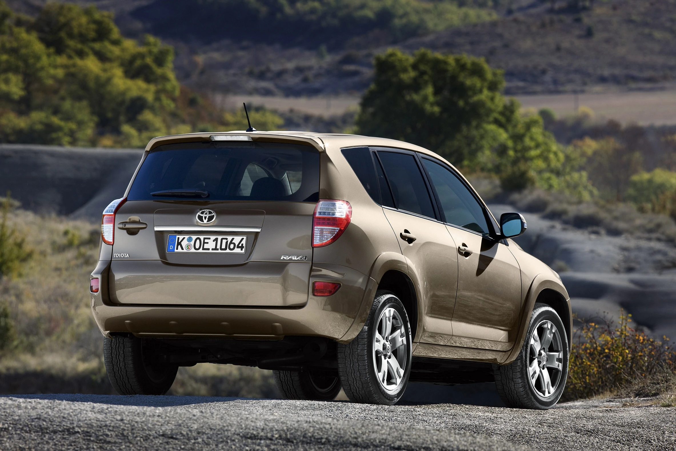 Beiger Toyota RAV4 parkt auf einem grauen, felsigen Gelände. Im Hintergrund sind Bäume und ein bewölkter Himmel zu sehen.